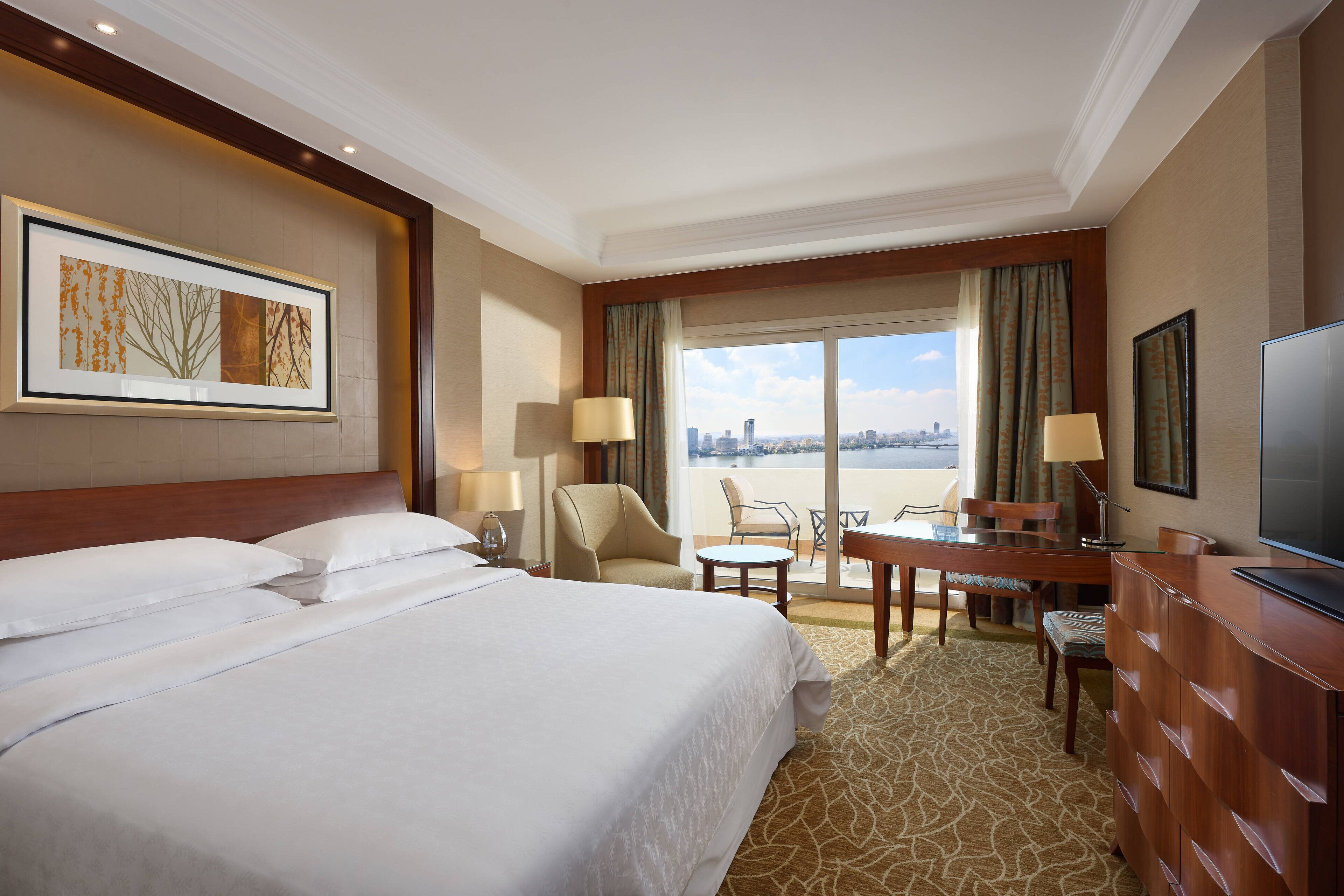 Habitación de hotel con una cama grande, un escritorio, una silla, un televisor y vistas a la ciudad desde la ventana.
