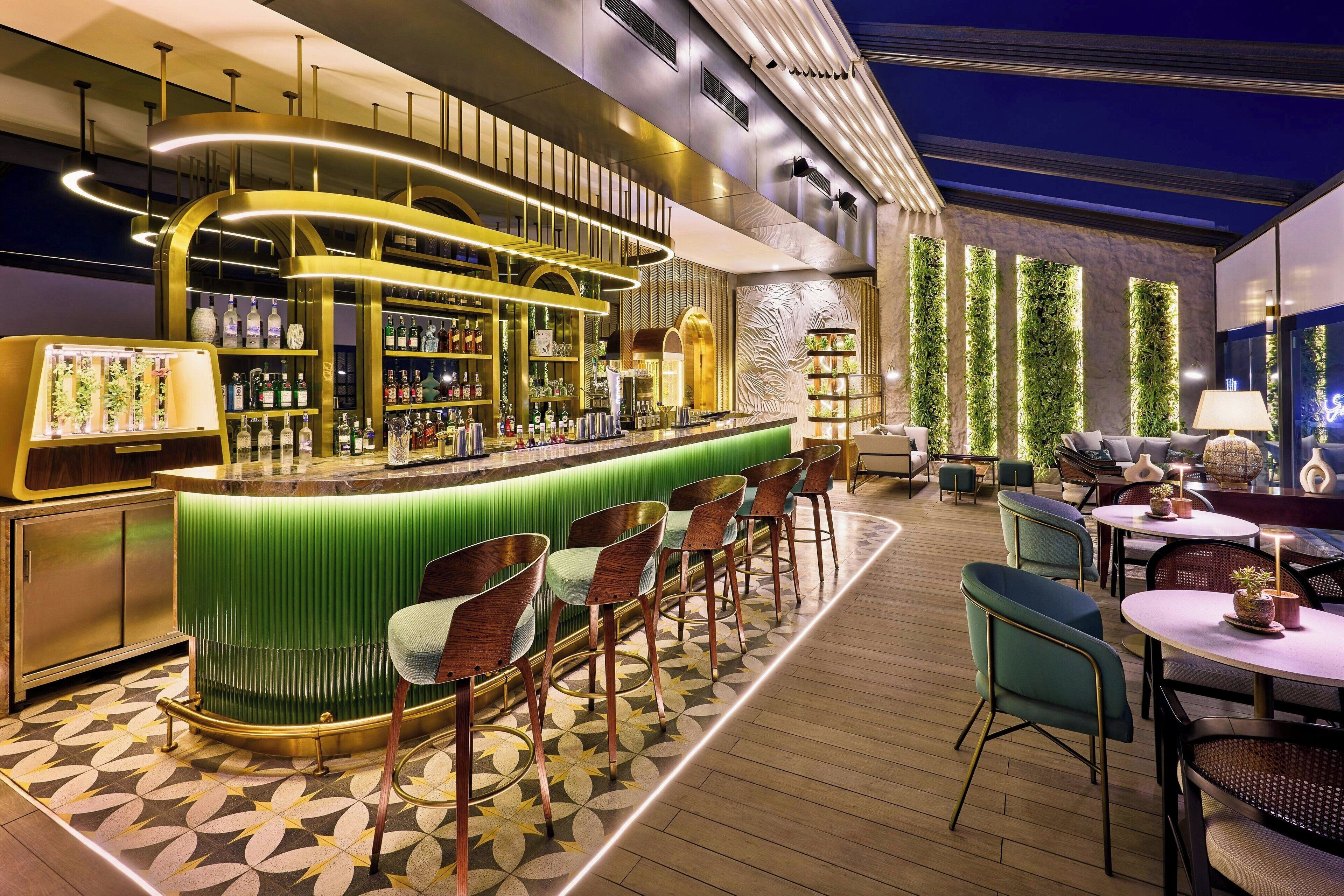 Eine Bar mit einem grünen Tresen, mehreren Barhocker und einer Auswahl an Flaschen. Es gibt auch mehrere Tische und Stühle zum Essen.