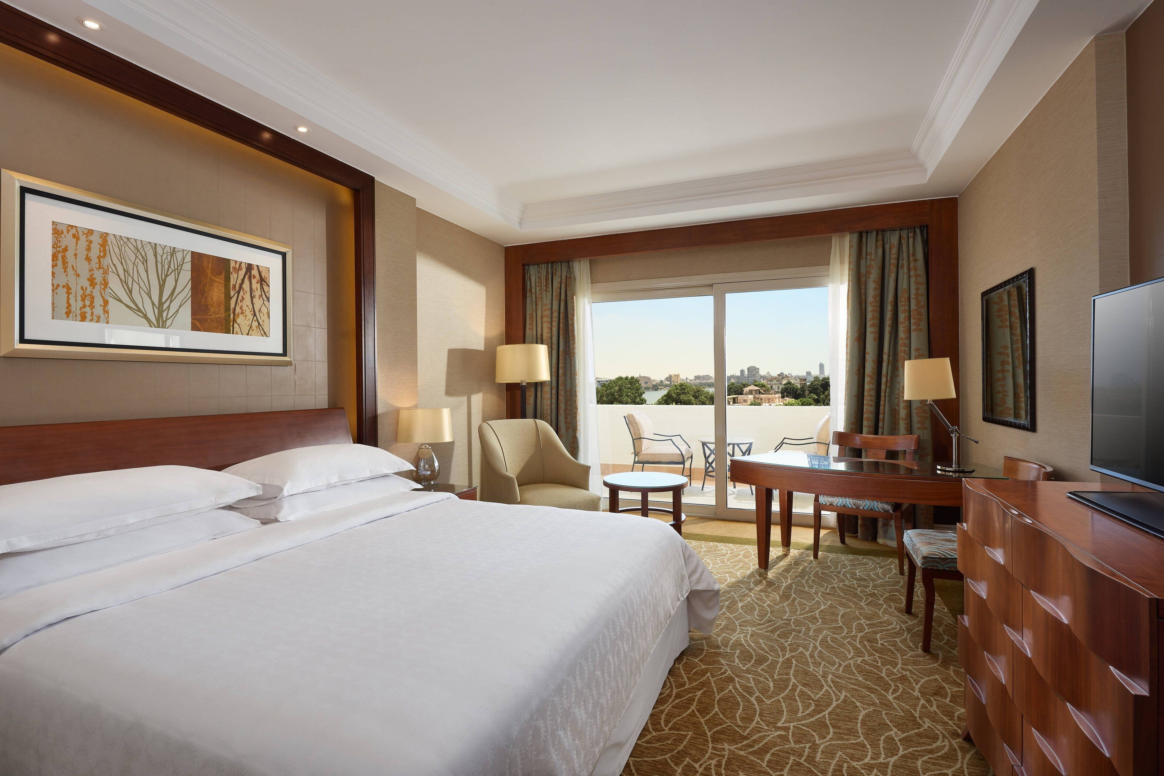 Habitación de hotel con una cama grande, un escritorio con silla, televisión y un balcón con vista a la ciudad.