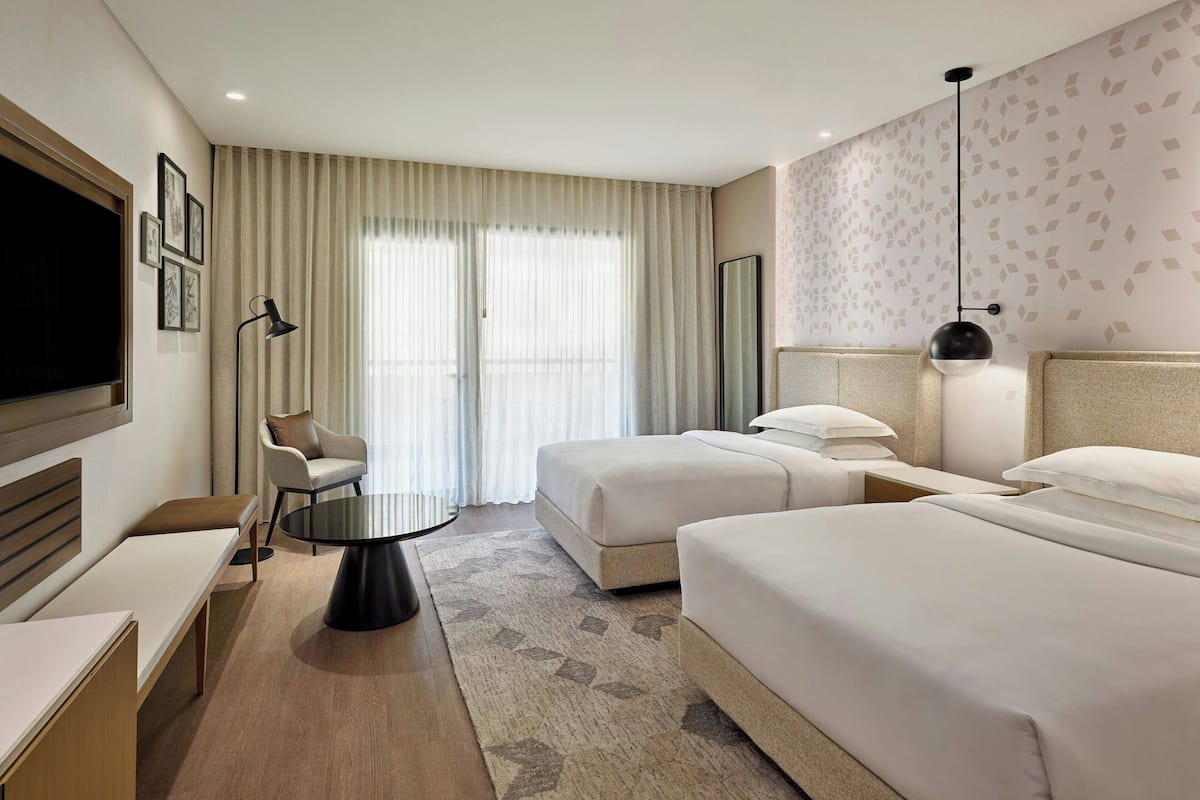 Habitación de hotel con dos camas, un escritorio, una silla, televisión y un ventanal con cortinas.