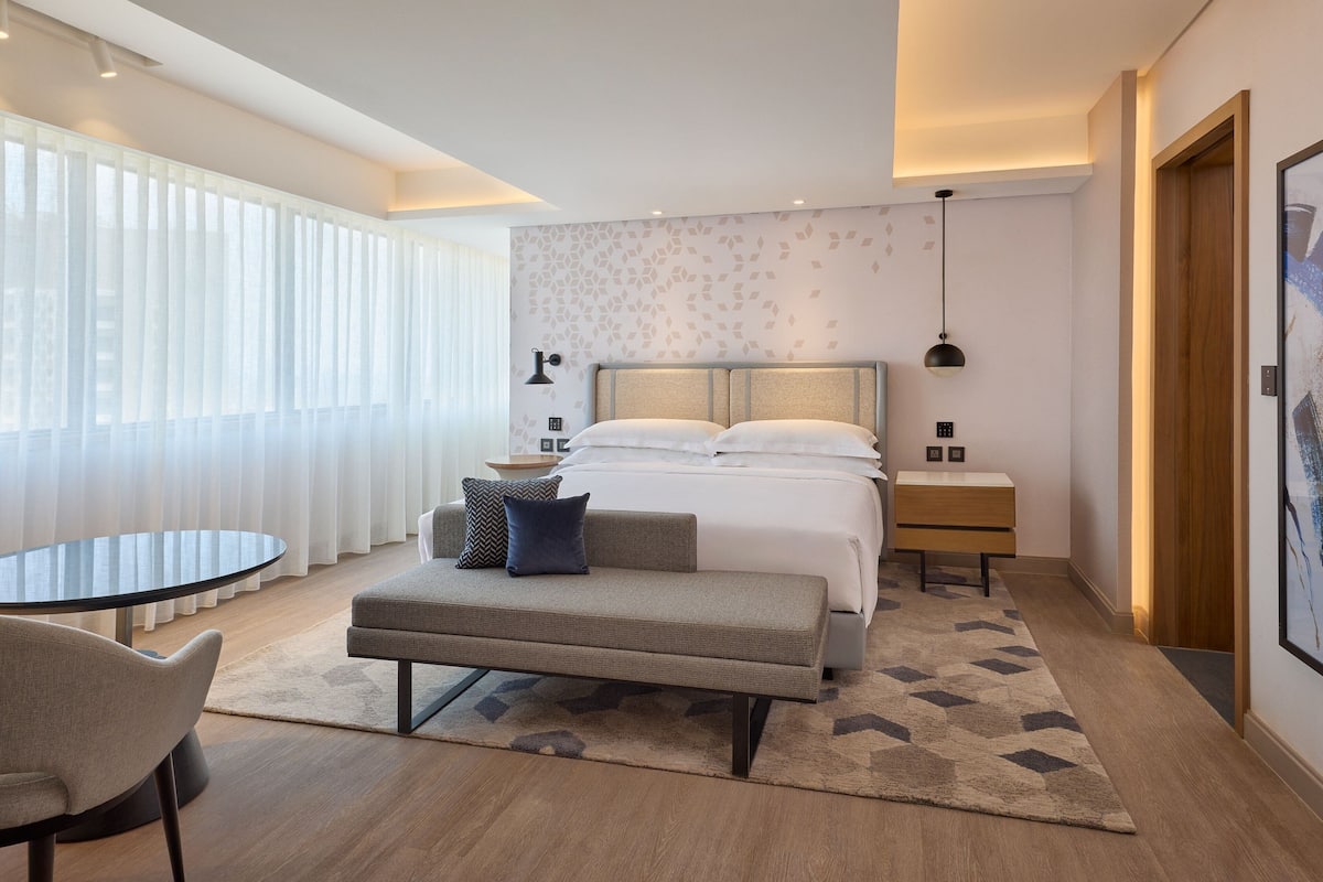 Una habitación de hotel moderna con una cama grande, un sofá pequeño, una mesa redonda y un ventanal con cortinas translúcidas.