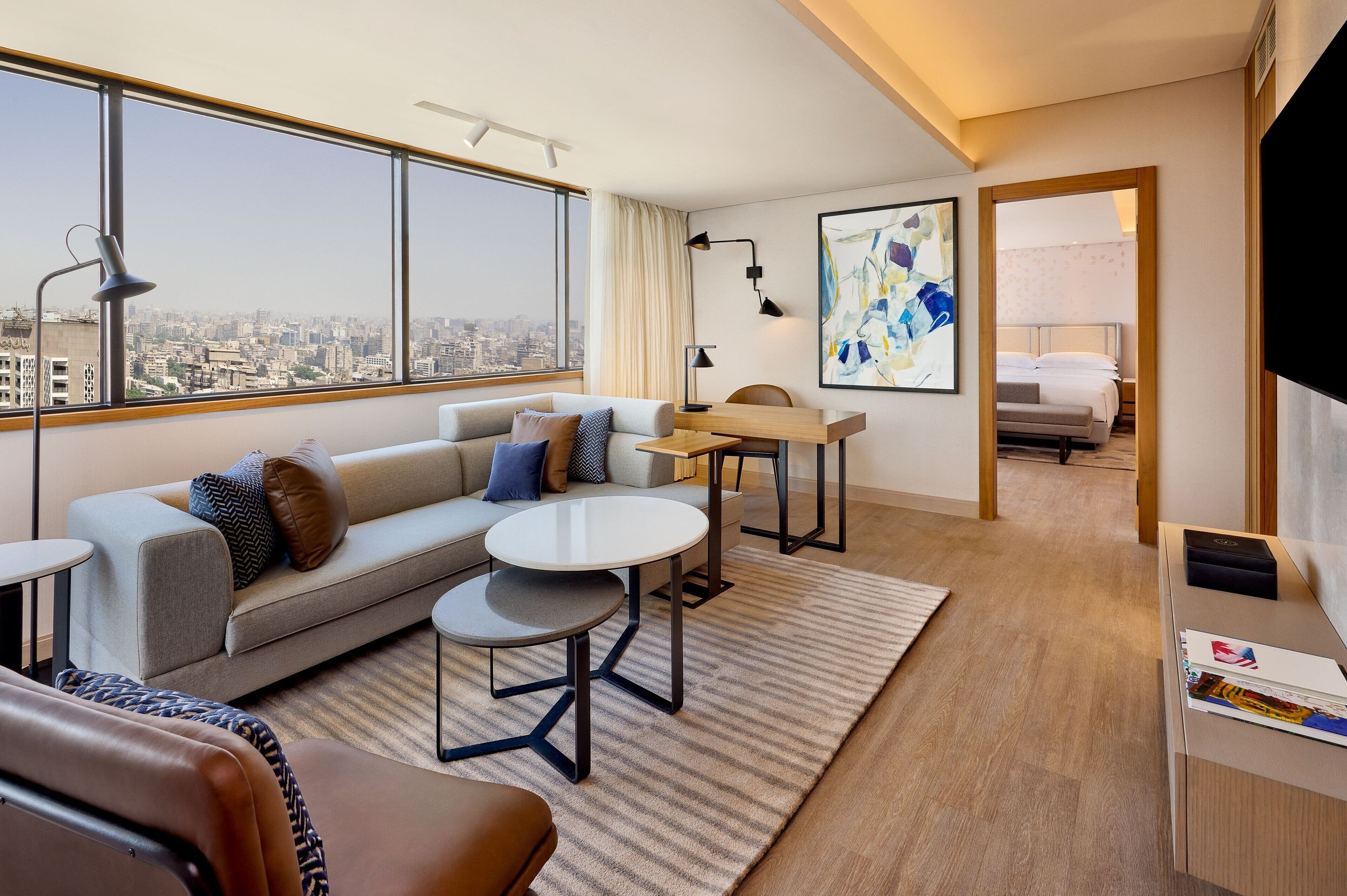 Una habitación de hotel moderna con un ventanal grande, un sofá, una mesita redonda pequeña, un escritorio y una cama al fondo.
