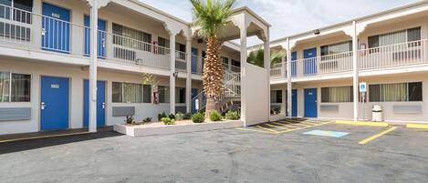 A multi-story motel with blue doors numbered 210 and 119, a central palm tree, and a parking area with marked spaces.