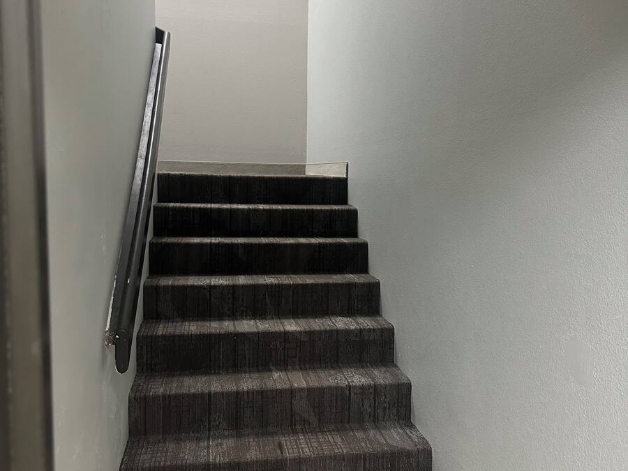 A staircase with dark carpeted steps and a metal handrail, leading to a landing with a white wall.