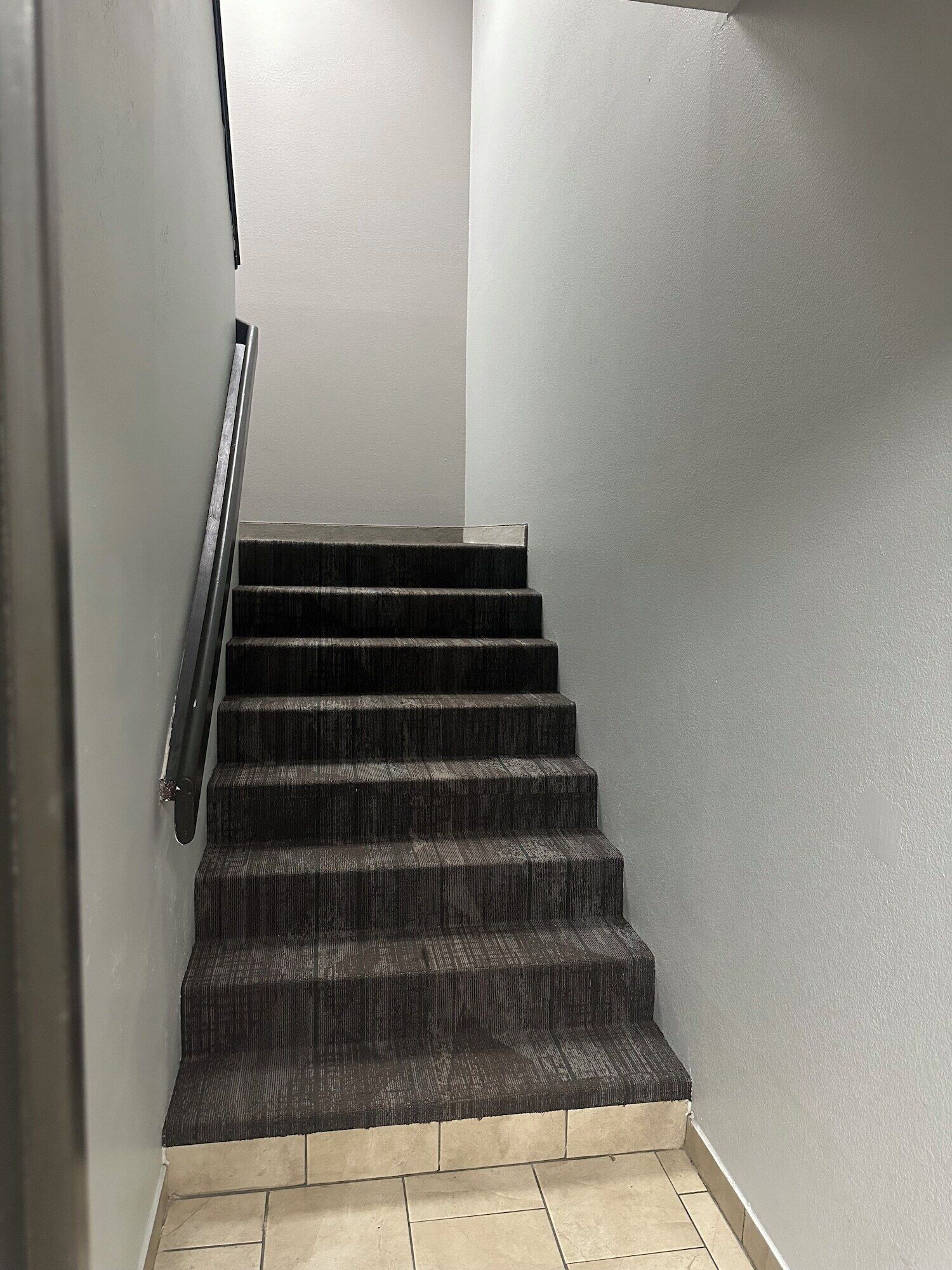 Egy lépcsőház sötét színű, szőnyeggel borított lépcsőfokkal és fém korláttal, amely egy fehér falú, pihenőhelyhez vezet.