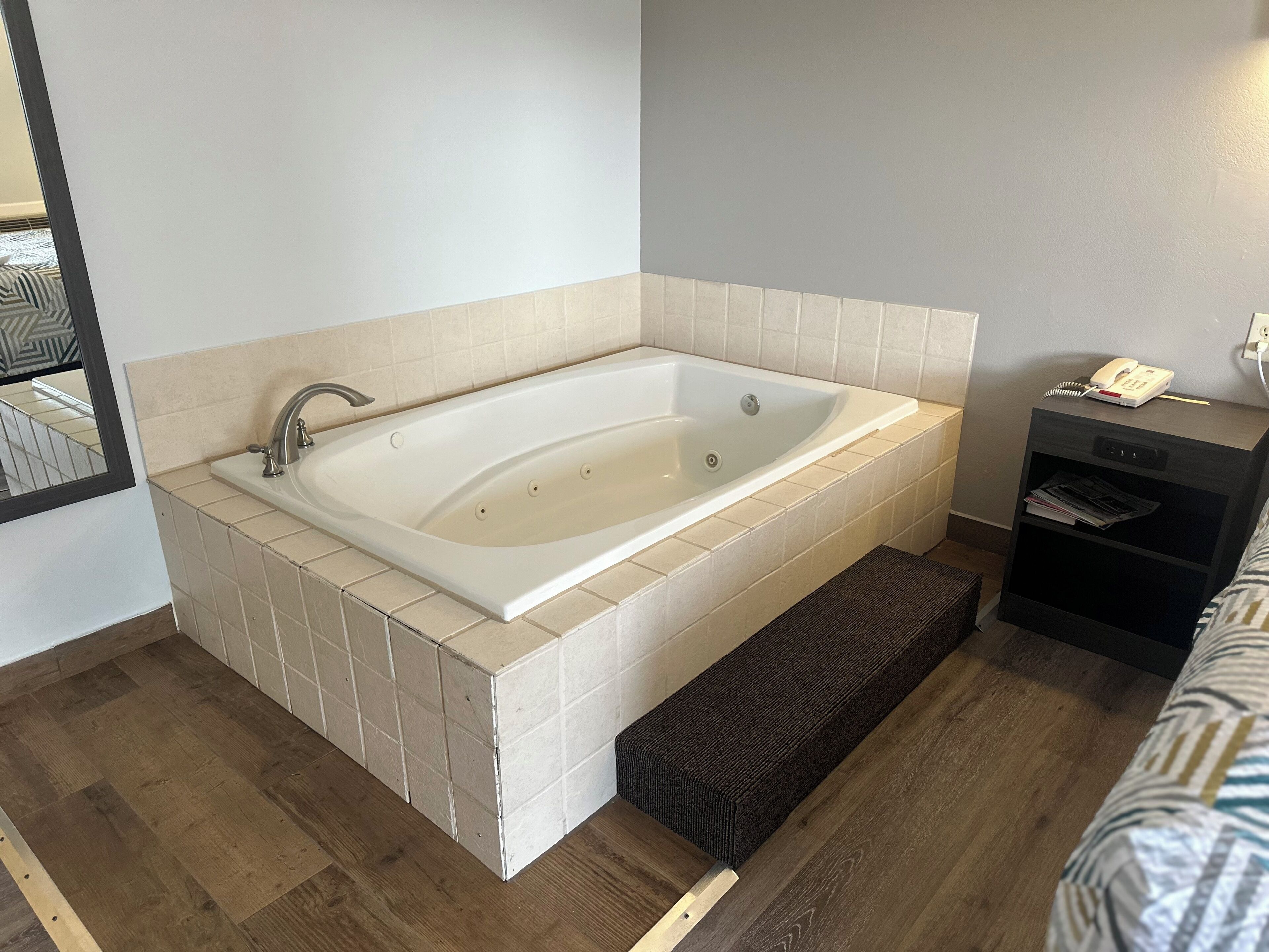 A modern bathroom with a freestanding bathtub, a small bedside table with a phone, and a mirror.