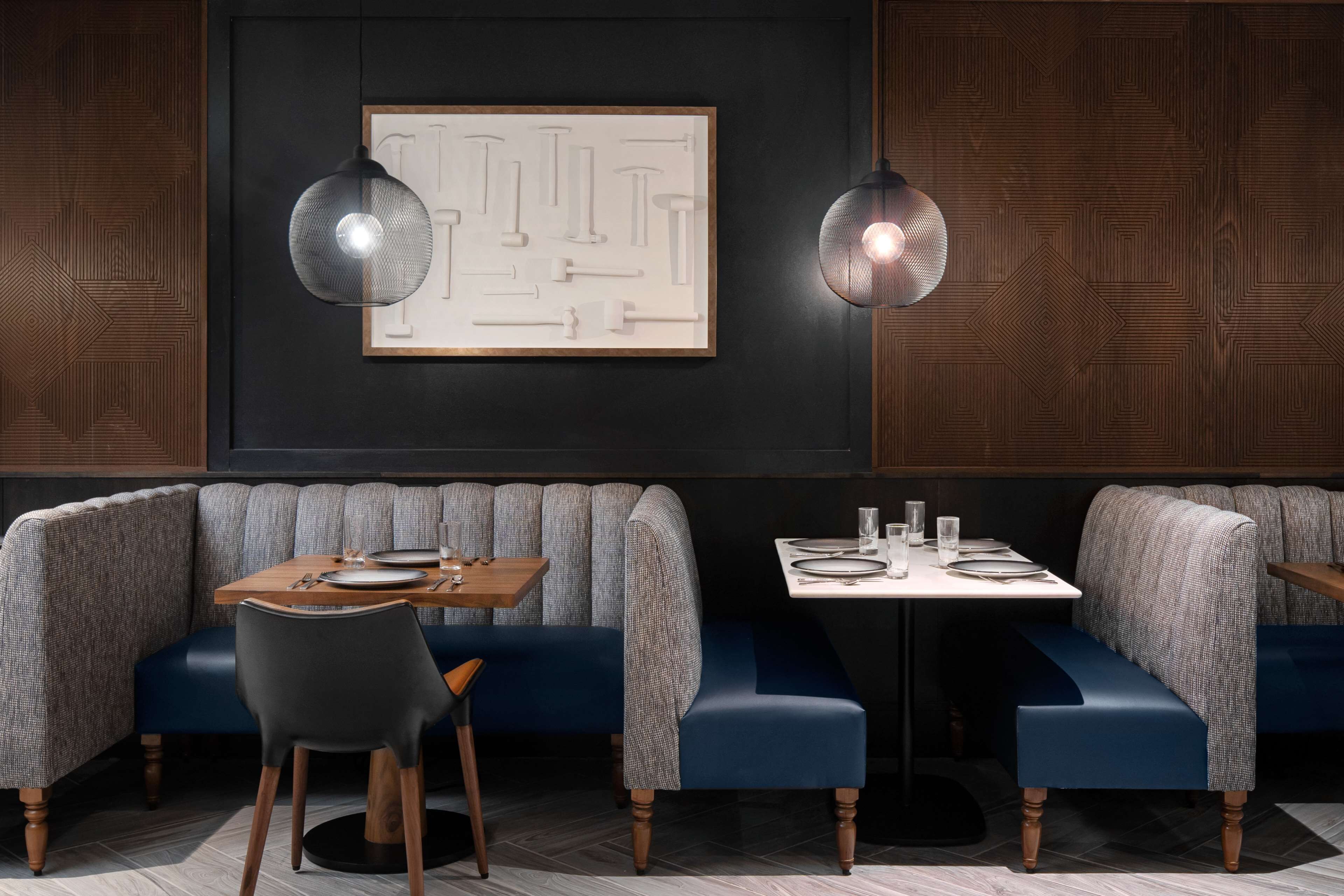 A restaurant interior with a dark wood panel wall, a framed abstract artwork, two pendant lights, a blue upholstered booth, a wooden table with a chair, and a white table with a glass.