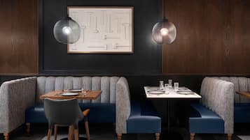 A restaurant interior with a dark wood panel wall, a framed abstract artwork, two pendant lights, a blue upholstered booth, a wooden table with a chair, and a white table with a glass.