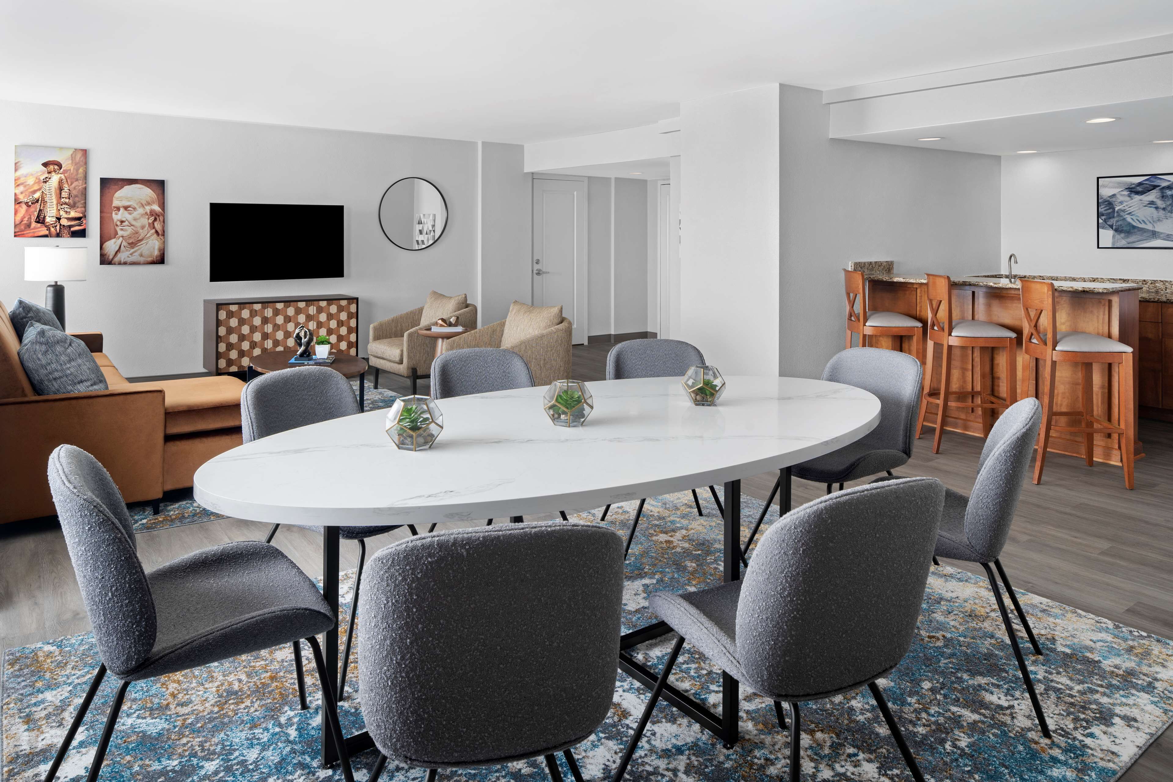 Un salon moderne avec une table ronde, des fauteuils à revêtement gris, un canapé et un téléviseur fixé au mur.