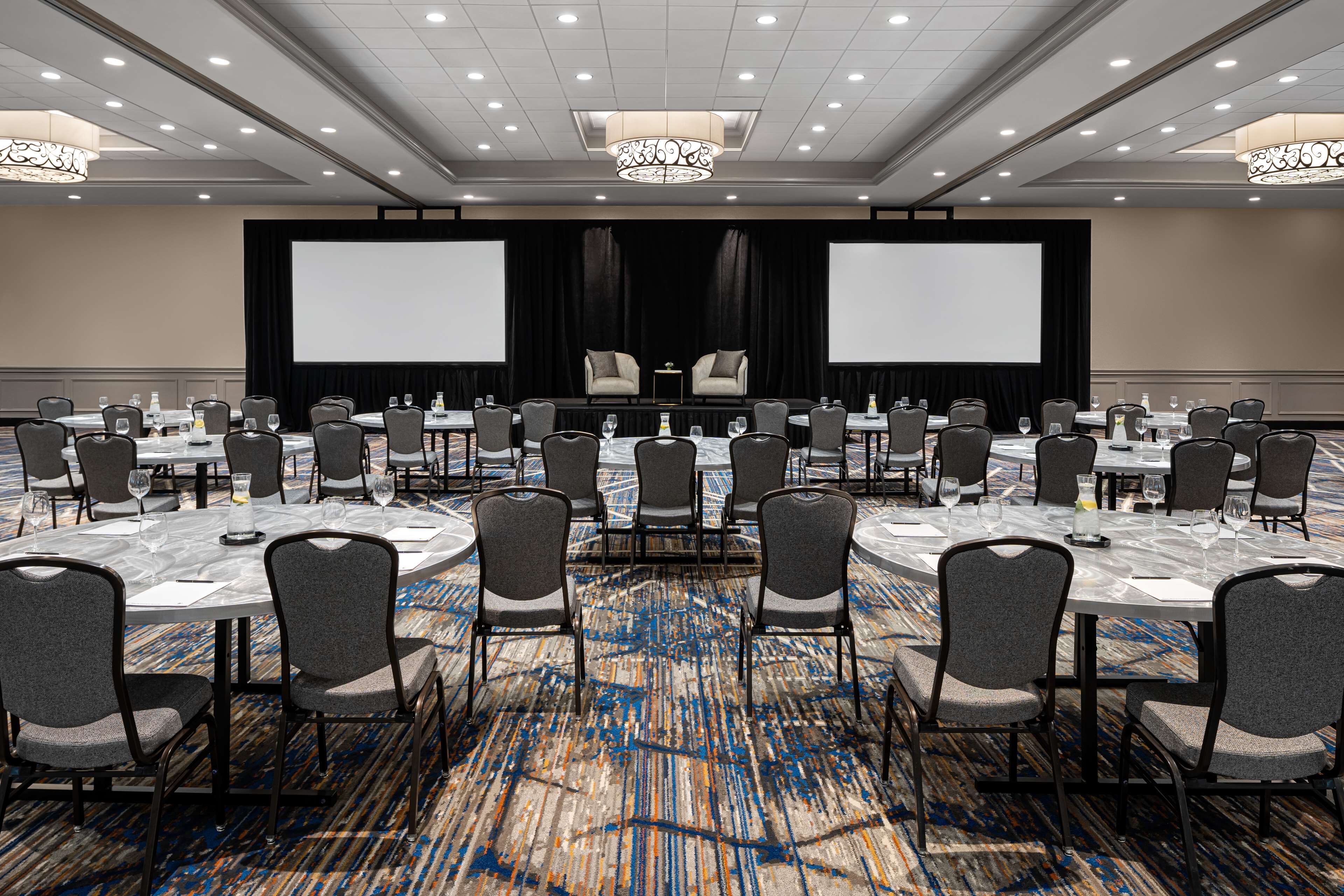 A conference room with multiple tables and chairs, a large screen, and a stage with curtains.