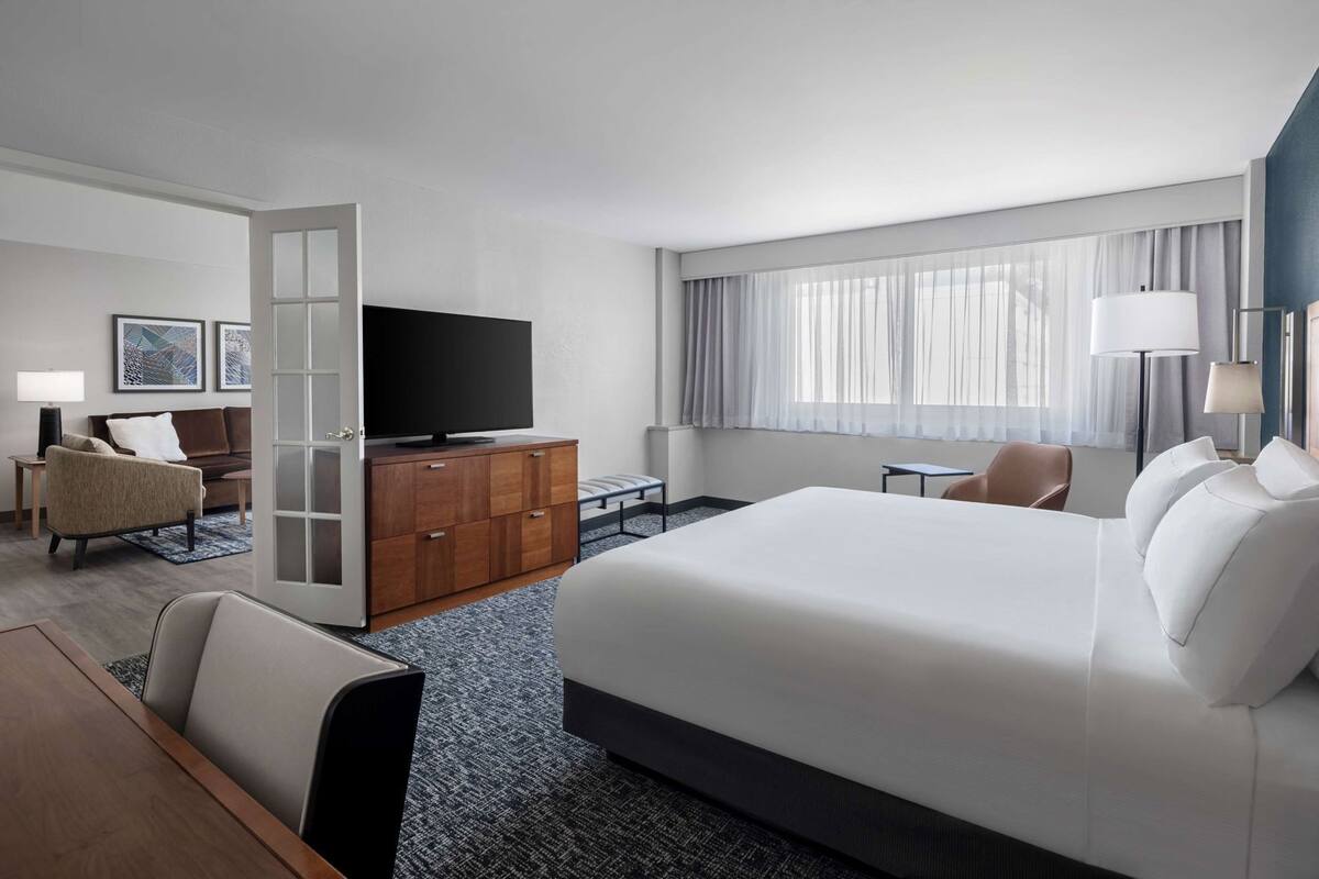 Ein Hotelzimmer mit einem großen Bett, einem Fernseher, einem Schreibtisch, einem Sessel und einem Kleiderschrank.