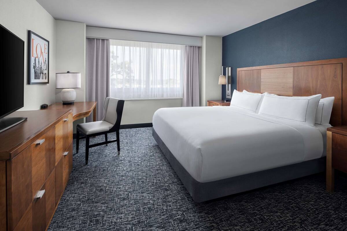 Ein Hotelzimmer mit einem großen Bett, einem Schreibtisch, einem Sessel, einem Fernseher und einem Fenster mit Vorhängen.