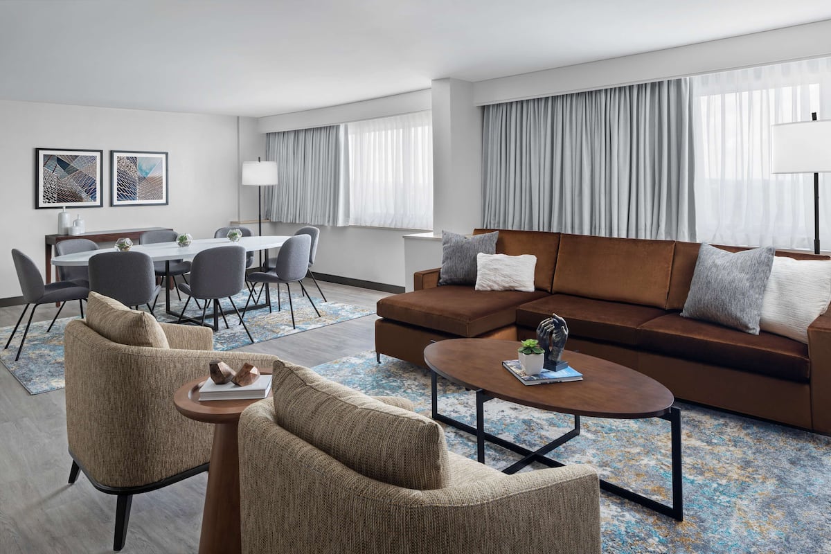 A modern hotel room with a living area, dining space, and large windows with curtains.