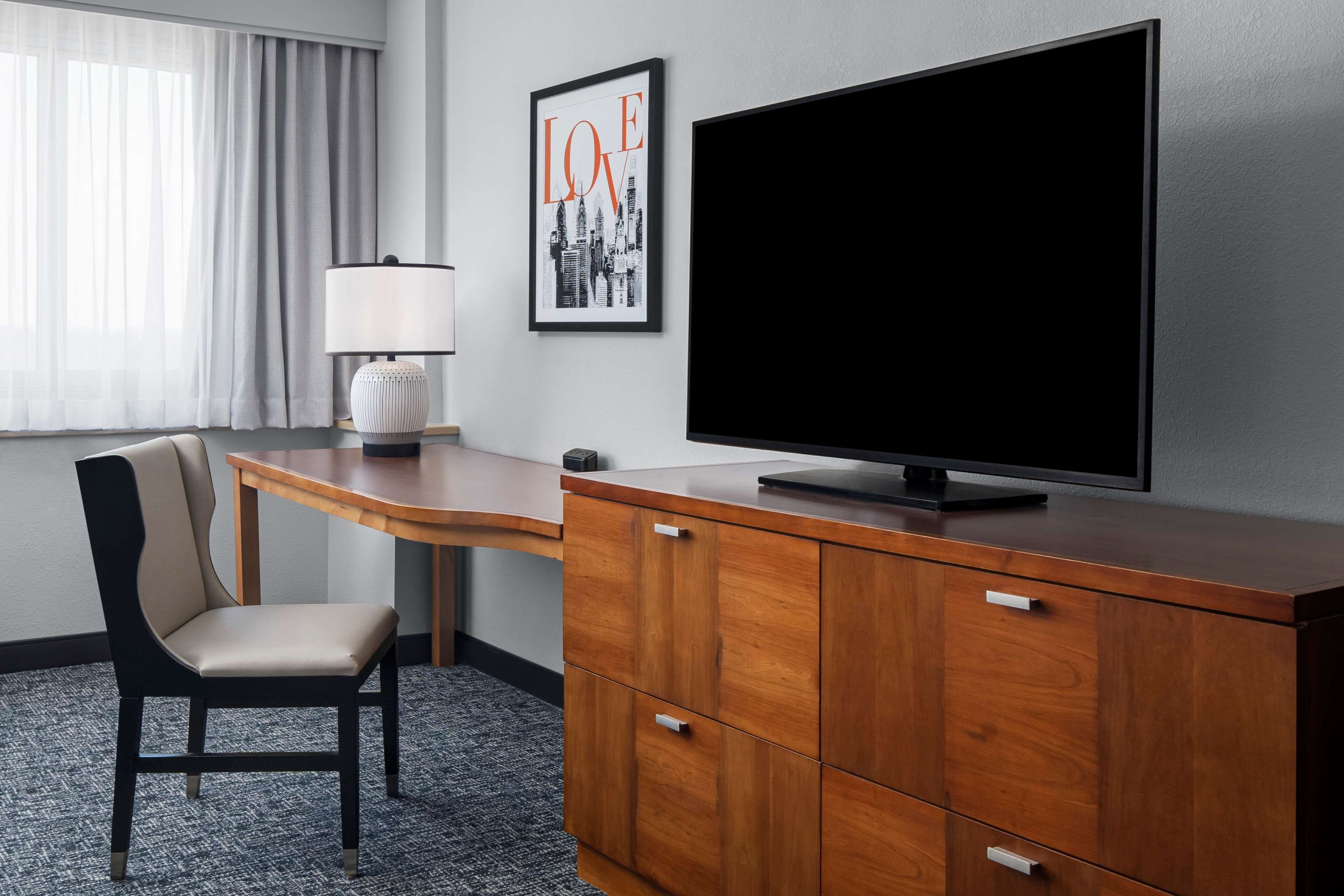 A hotel room with a wooden desk, a television, a chair, and a framed picture on the wall.