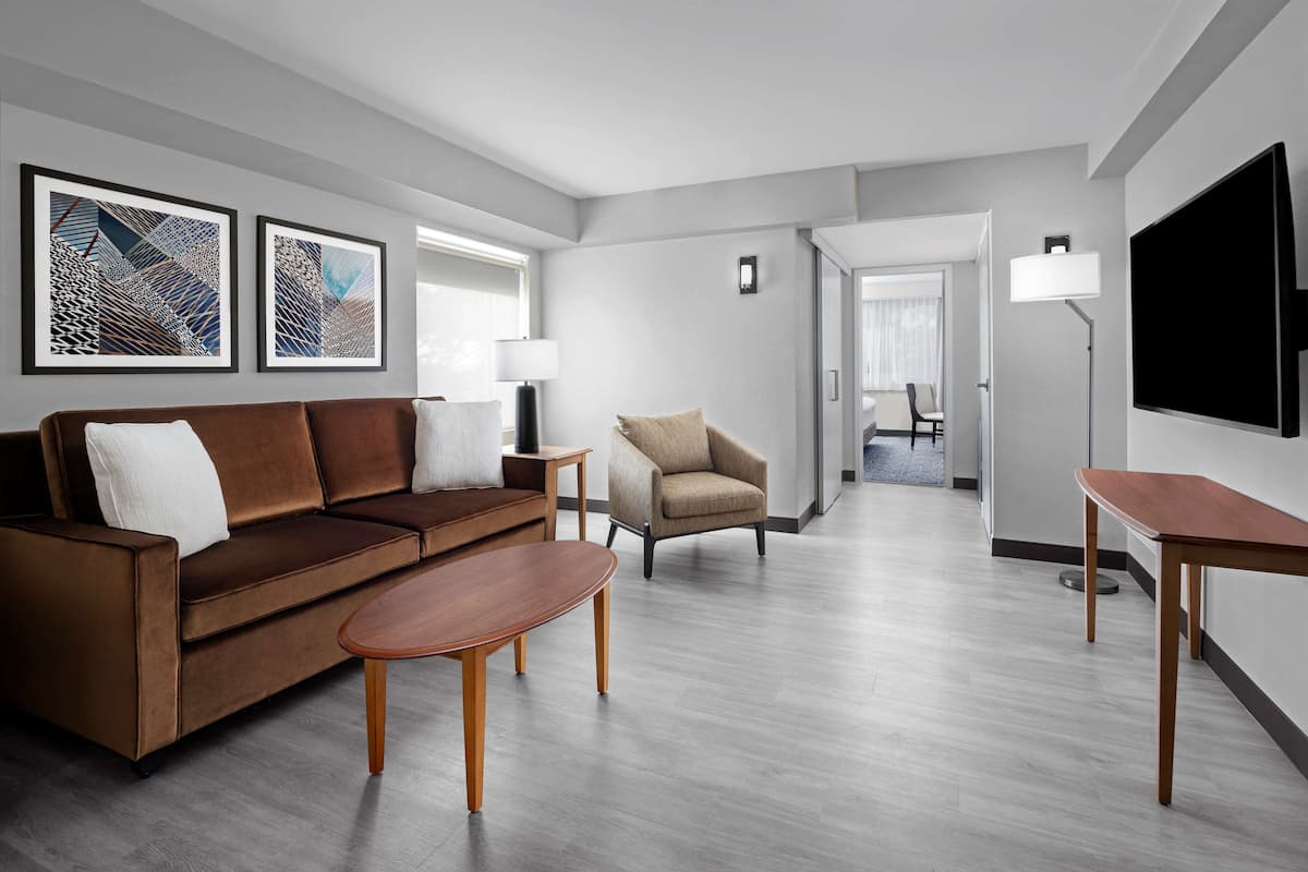 A modern living room with a brown sofa, a beige armchair, and a wooden coffee table. There are framed artworks on the wall and a flat-screen TV mounted on the wall.