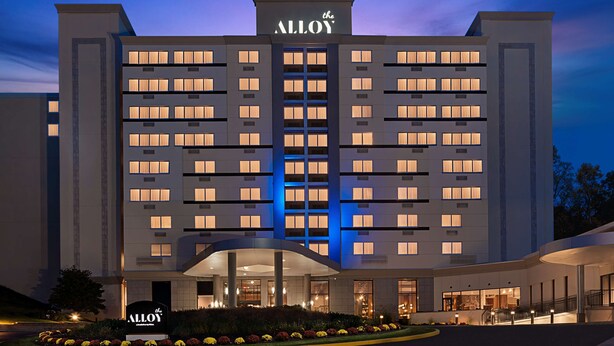 Un hotel de varios pisos con un llamativo letrero 'Alloy', un acceso cubierto y una zona ajardinada con flores.