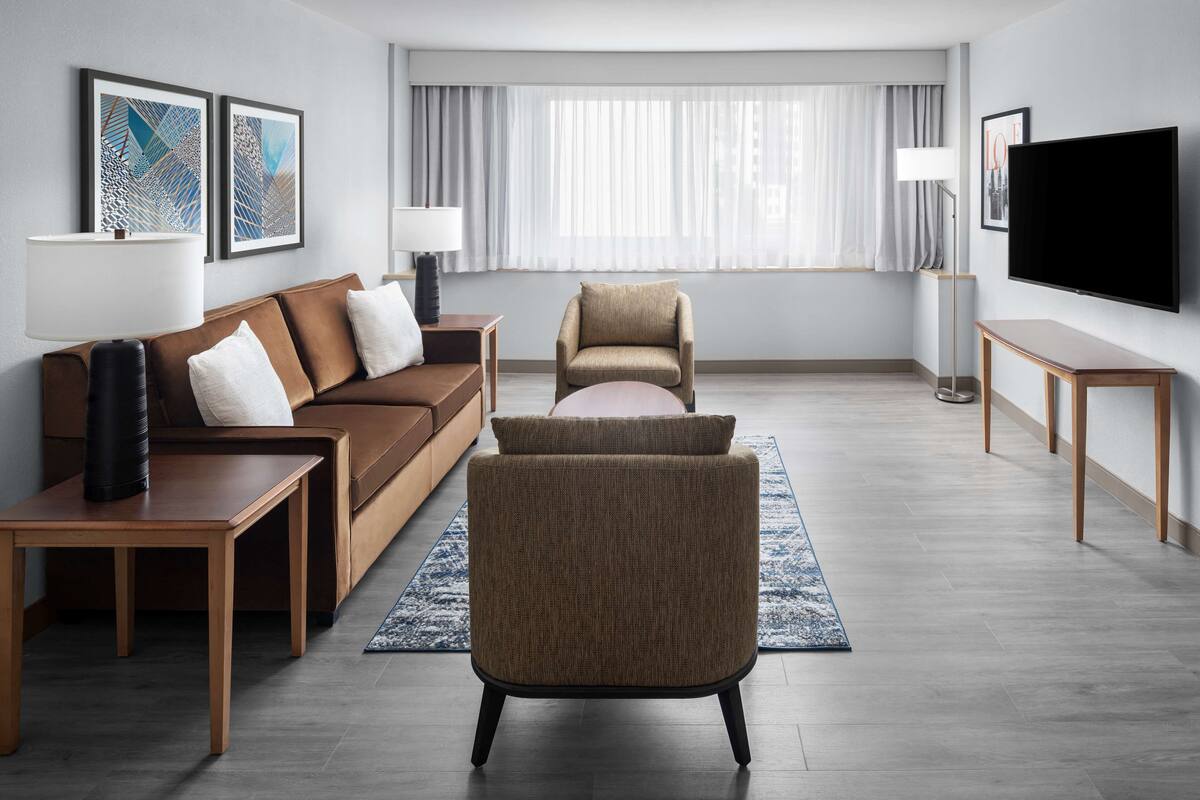 A modern living room with a brown sofa, a beige armchair, and a wooden coffee table. There is a flat-screen TV mounted on the wall, a floor lamp, and two framed artworks on the wall.