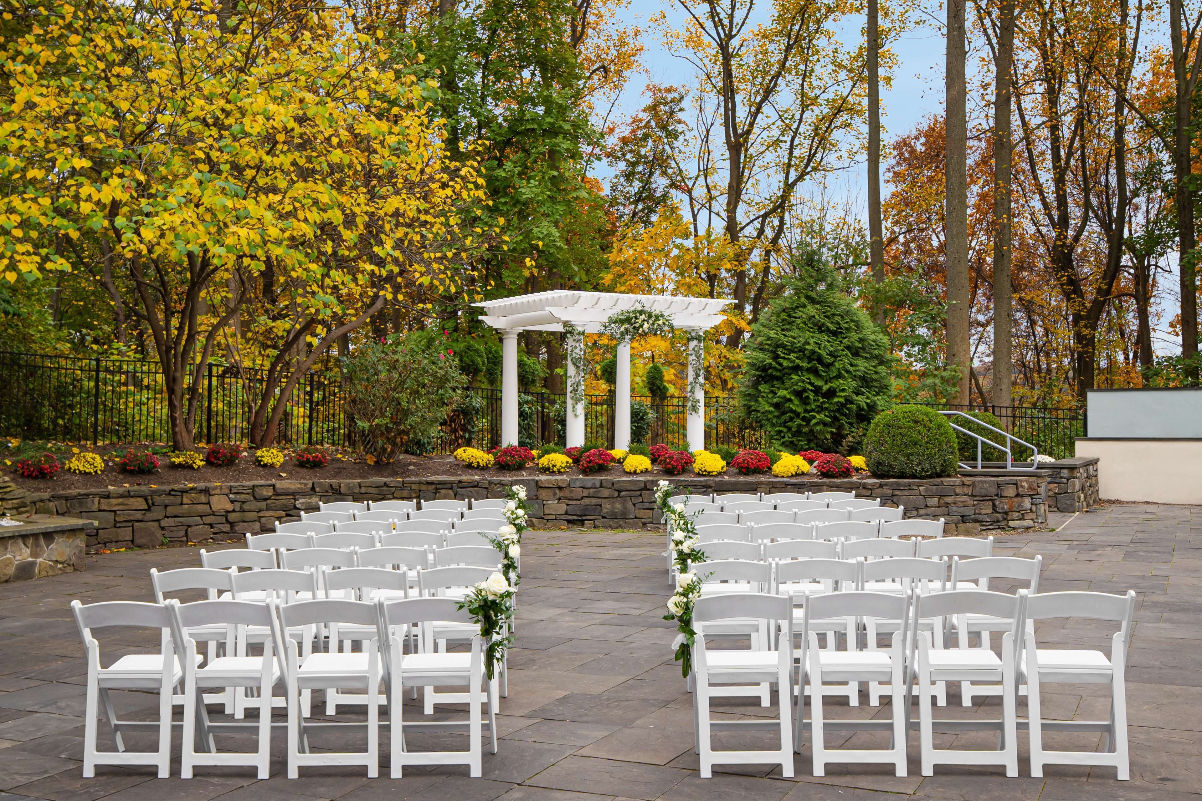 An outdoor wedding venue with white chairs arranged in rows, a white archway, and a stone wall with colorful flowers.