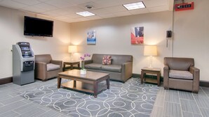 A hotel lobby with a flat-screen TV, an ATM, a sofa, two armchairs, a coffee table, and a patterned carpet.
