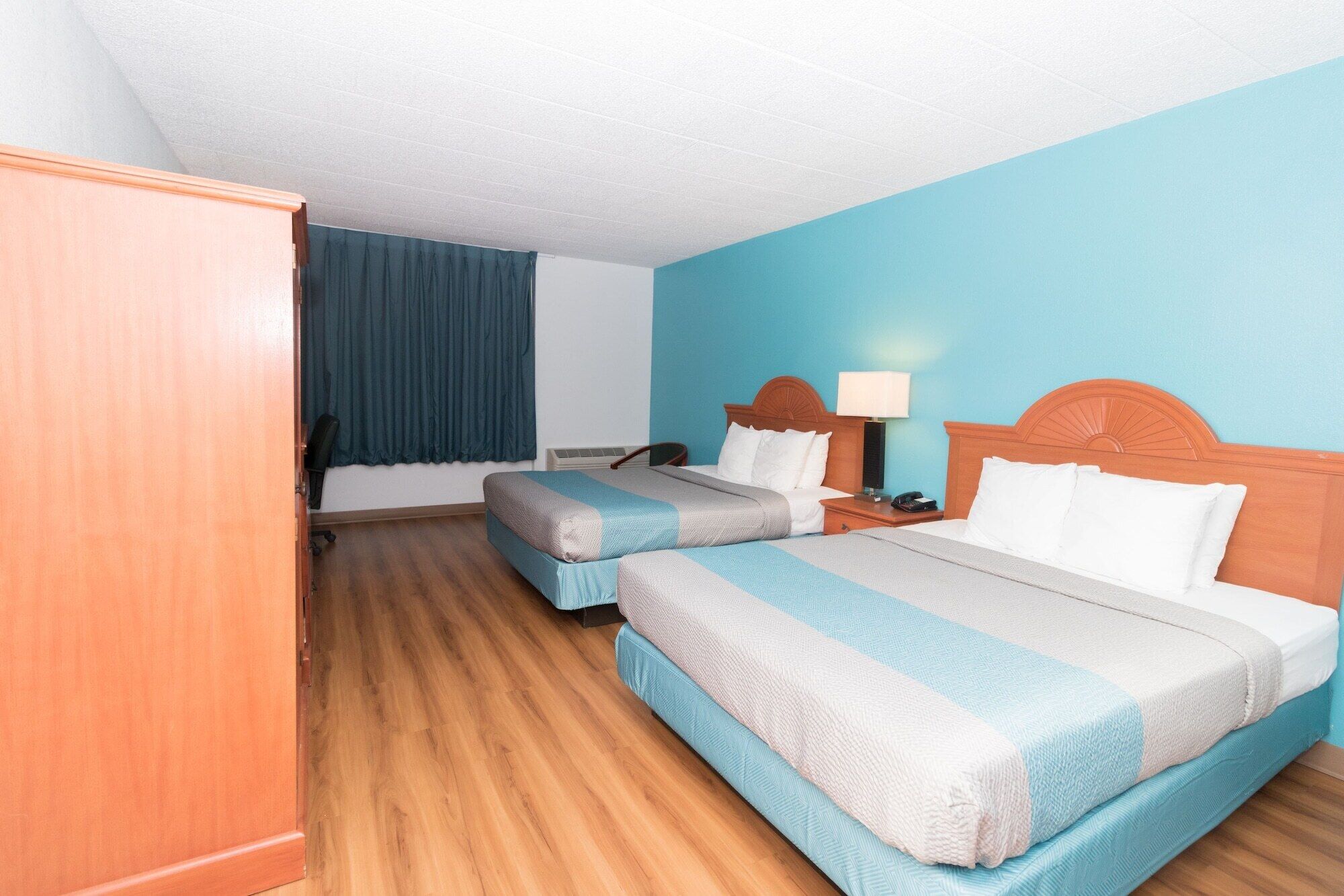 A hotel room with two beds, a wooden wardrobe, a desk with a lamp, and a window with curtains.