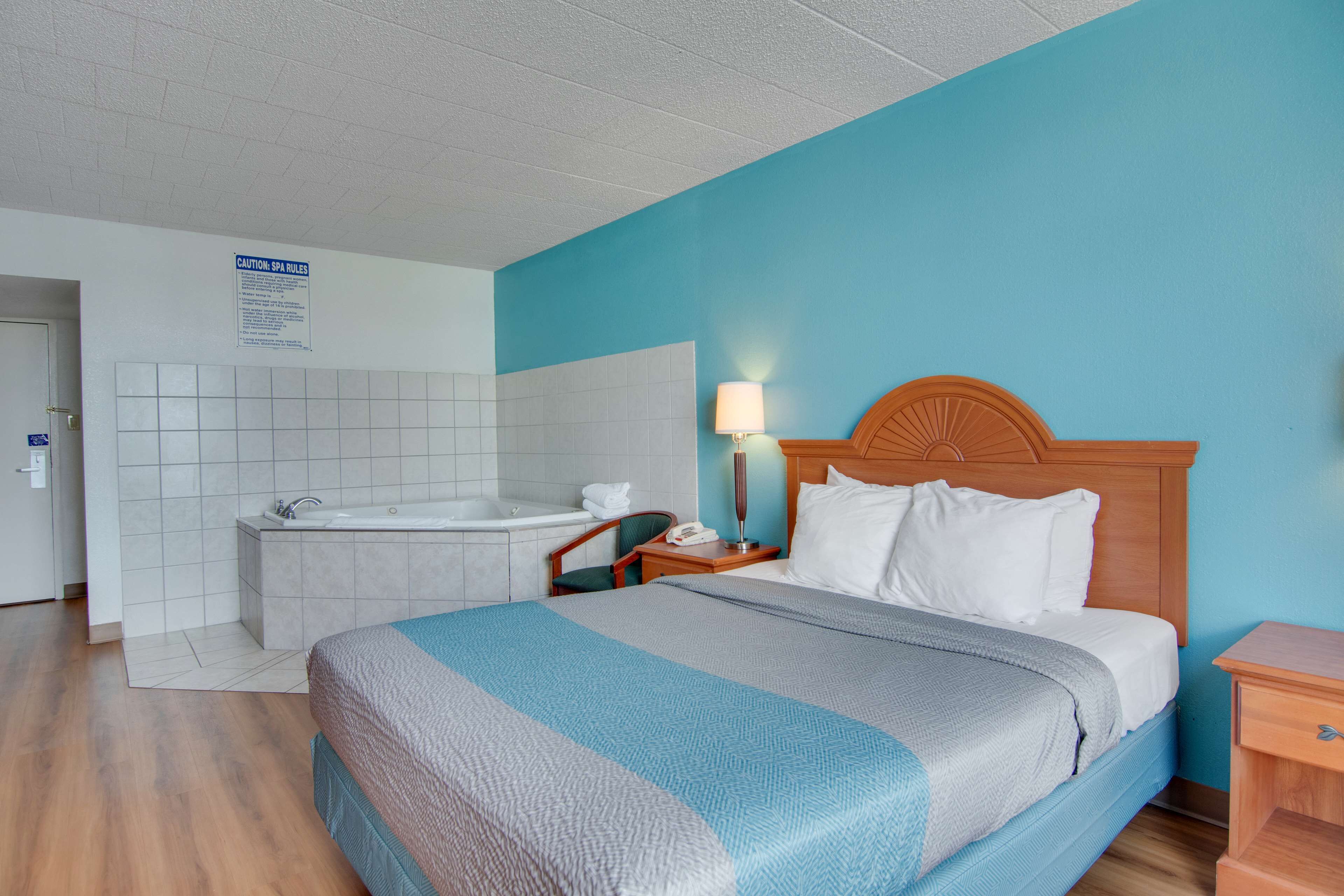 Habitación de hotel con una cama grande, cabecera de madera, mesita de noche con lámpara, baño con bañera y un cartel que indica las normas del spa.