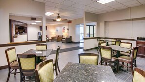 A hotel dining area with multiple tables and chairs, a kitchen in the background, and a ceiling fan.
