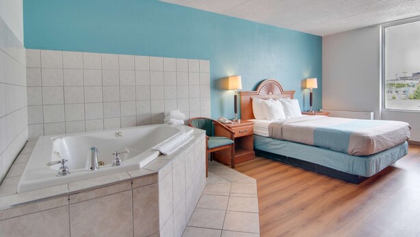 A modern hotel room with a large bathtub, a bed with a blue and white bedspread, a wooden nightstand with lamps, and a view of the city through a large window.