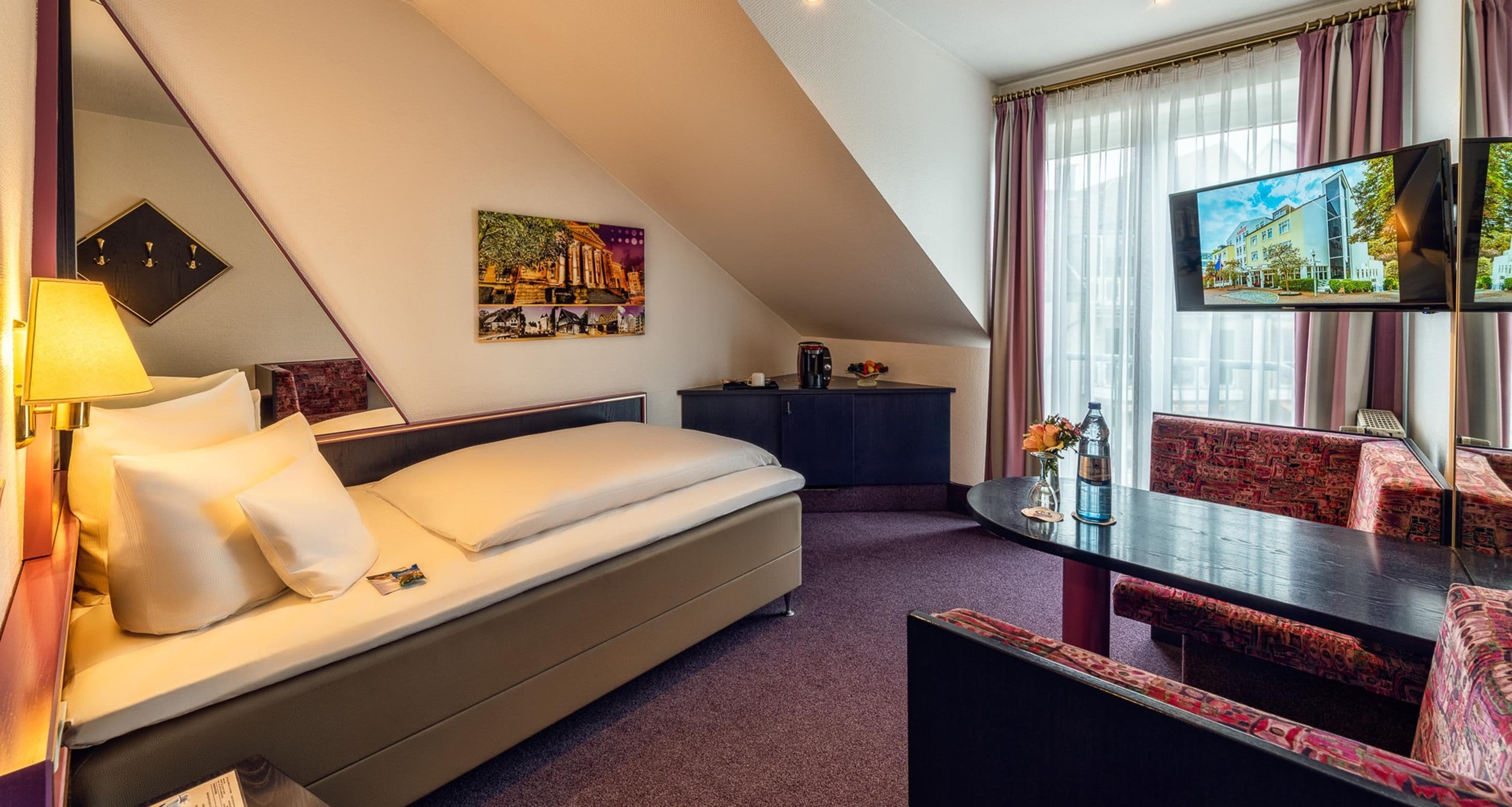 A hotel room with a bed, a desk, a chair, a television, and a painting on the wall.
