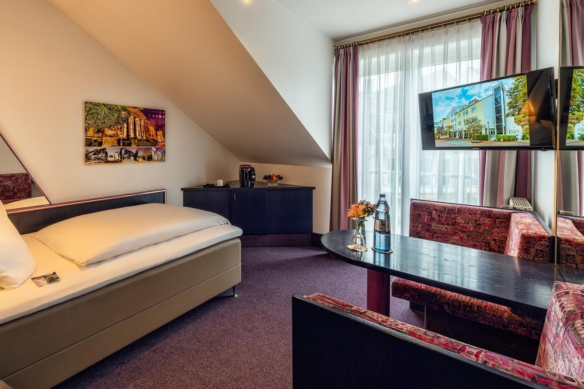 A hotel room with a bed, a desk, a chair, a television, and a painting on the wall.