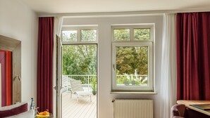 A hotel room with a bed, a desk, a chair, a window, and a balcony with a view of trees.