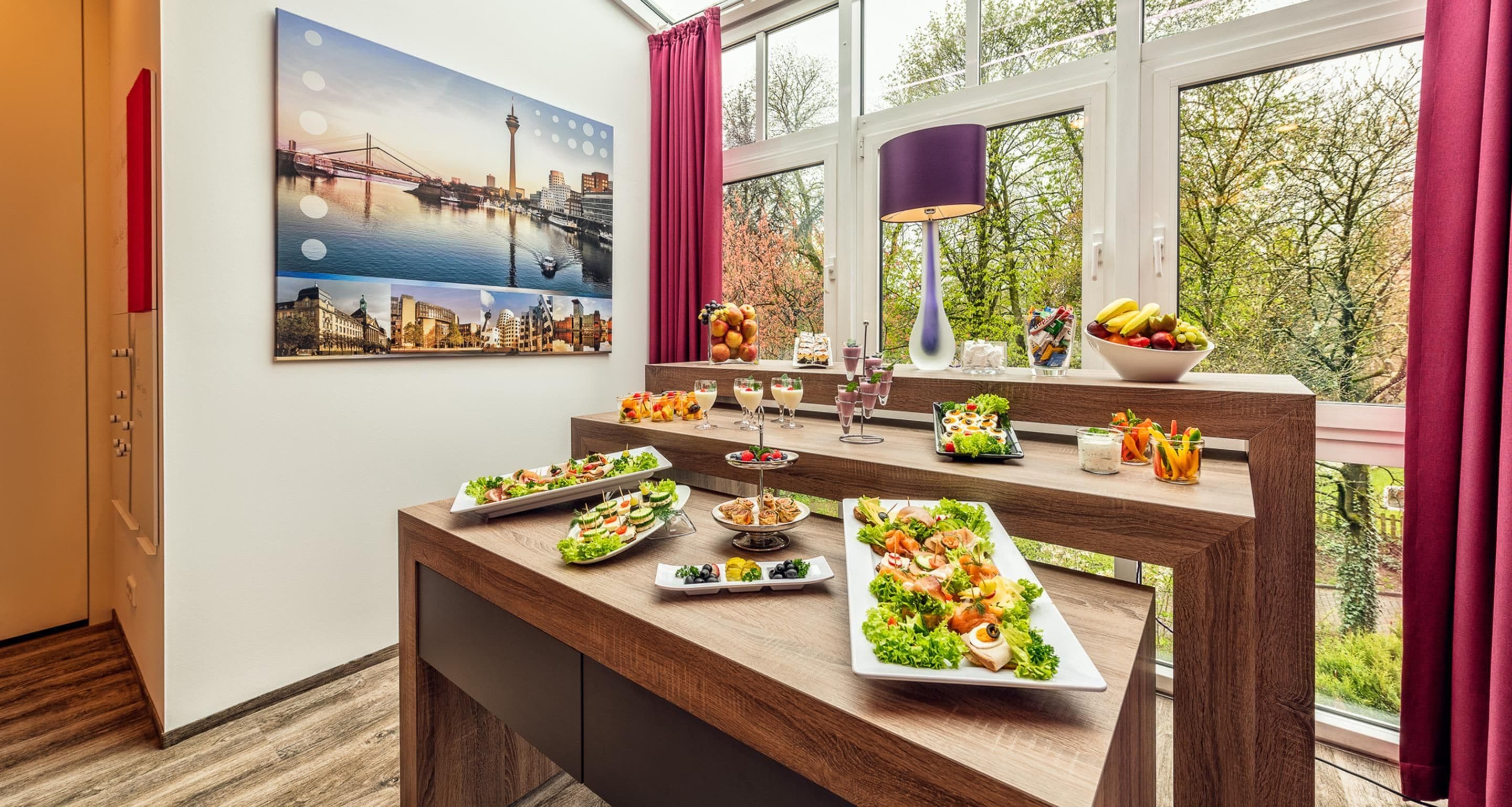 Un comedor con vistas a un paisaje urbano a través de amplios ventanales, una mesa de madera con diversas opciones de comida y un cuadro con un paisaje urbano en la pared.