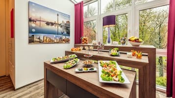 Un comedor con vistas a un paisaje urbano a través de amplios ventanales, una mesa de madera con diversas opciones de comida y un cuadro con un paisaje urbano en la pared.