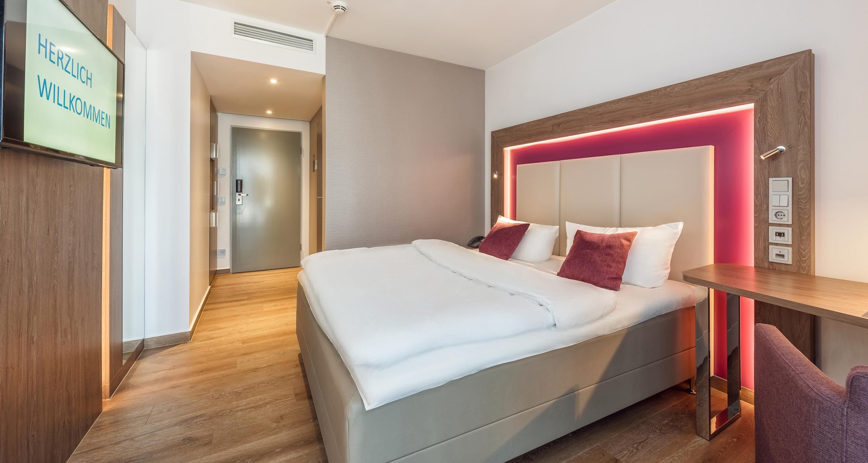 A modern hotel room with a large bed, bedside table, and a wall-mounted TV displaying 'HERZLICH WELCOME'.