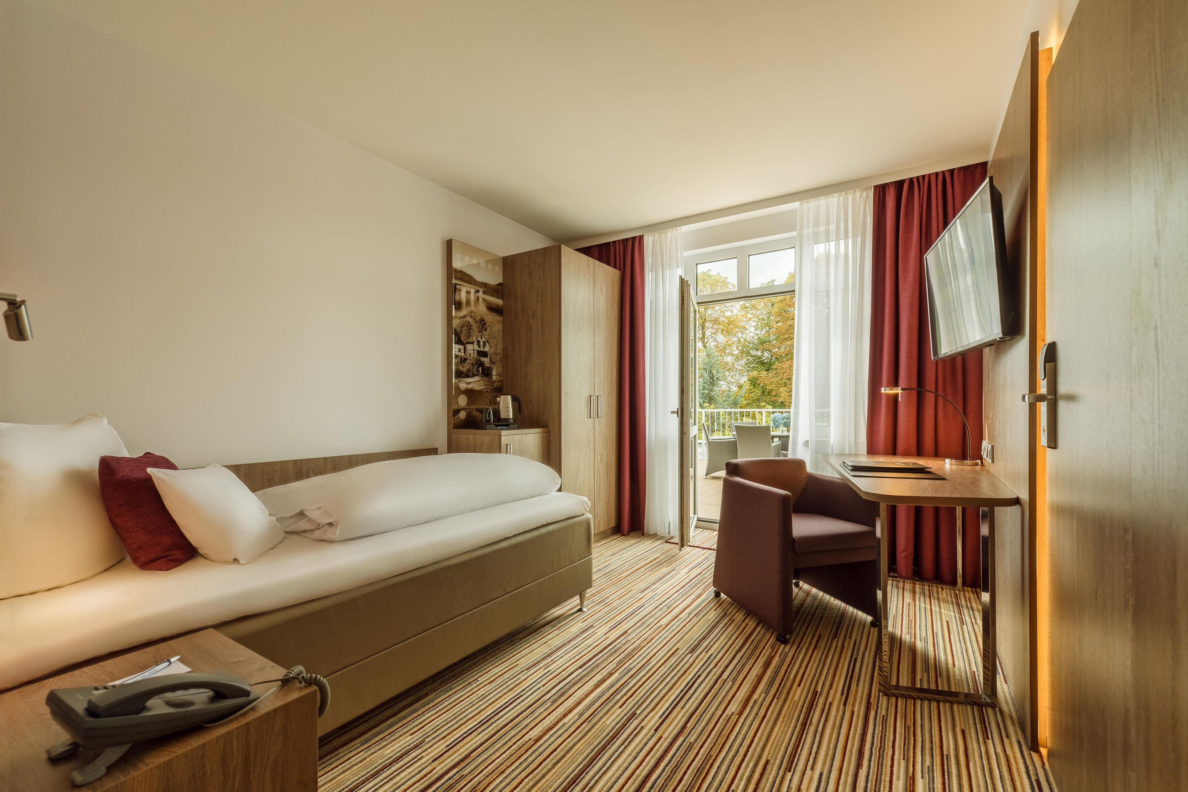 Habitación de hotel con sofá cama, escritorio con silla, televisión y una ventana grande con cortinas.