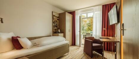 Habitación de hotel con sofá cama, escritorio con silla, televisión y una ventana grande con cortinas.