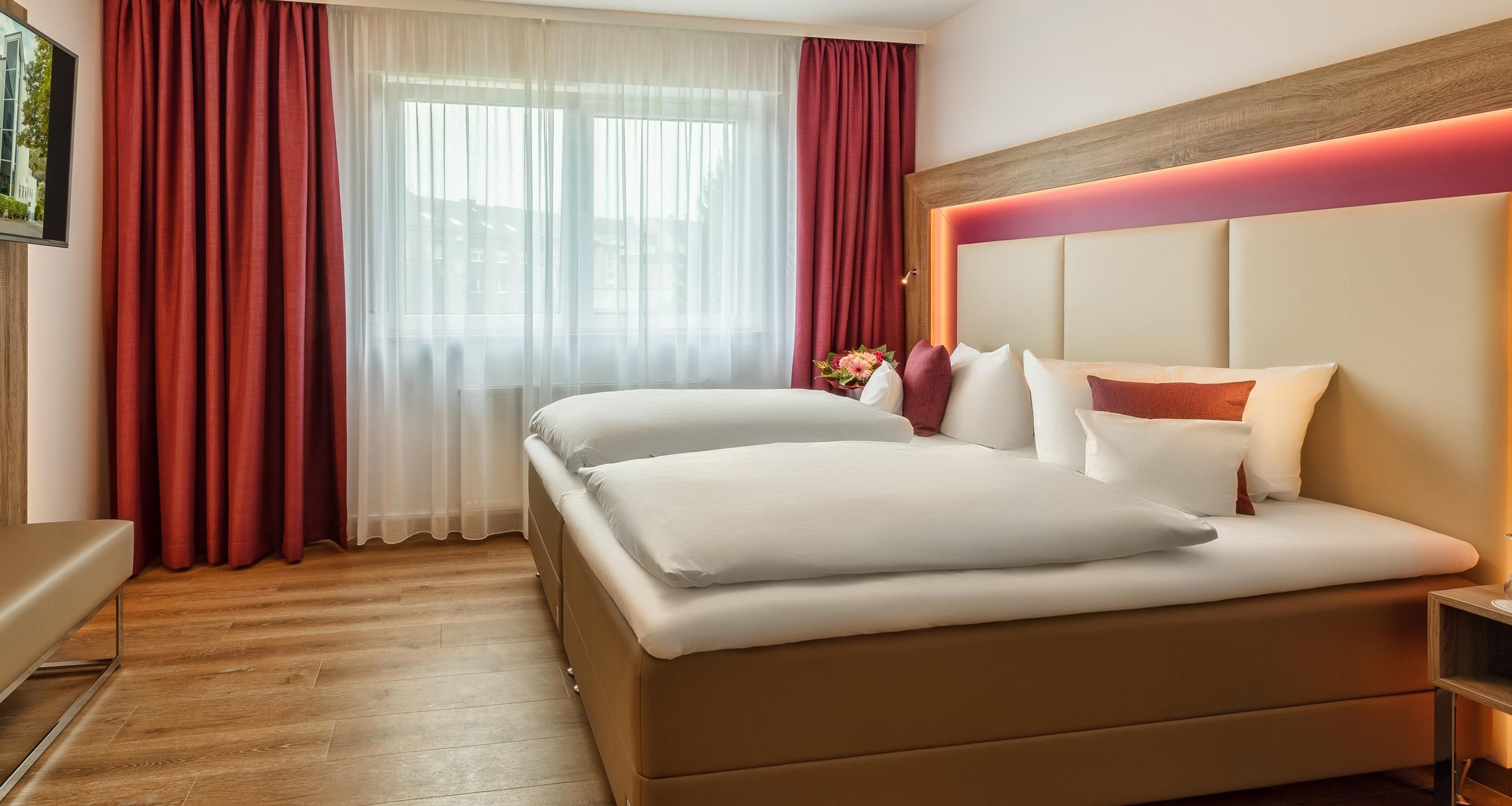 Habitación de hotel con cama, mesita de noche, cabecera de madera y ventana con cortinas translúcidas.