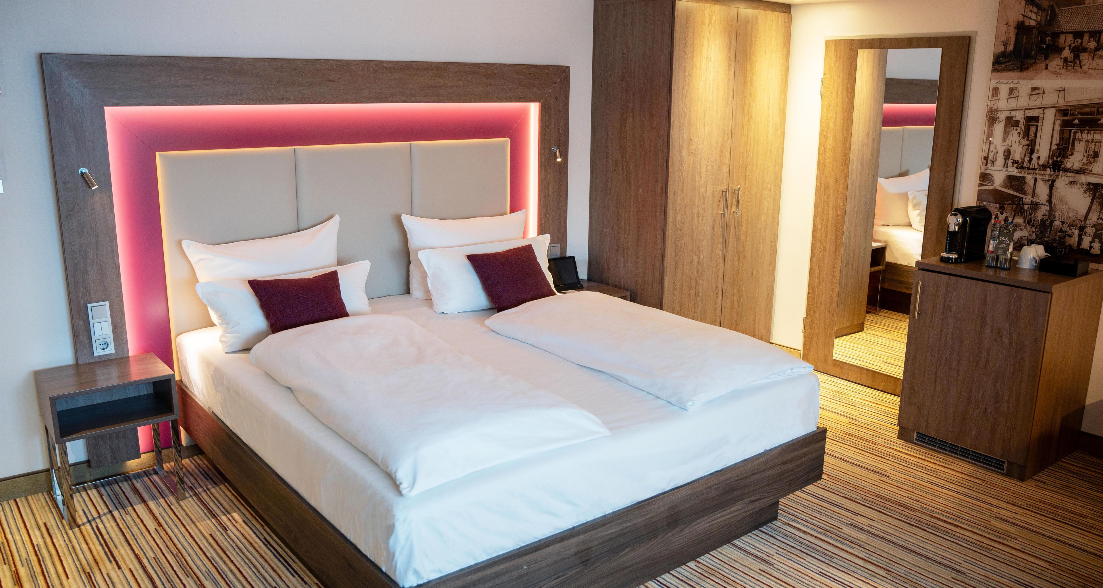 A modern hotel room with a large bed, a wooden headboard with integrated lighting, a nightstand, a wardrobe, and a small desk with a coffee machine.