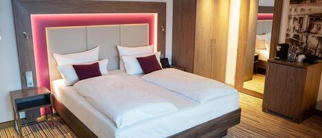 Una habitación de hotel moderna con una cama grande, cabecero de madera con iluminación integrada, una mesita de noche, un armario y un escritorio pequeño con una cafetera.