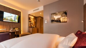 A hotel room with a bed, a television mounted on the wall, a painting of a building, and a kitchenette area.