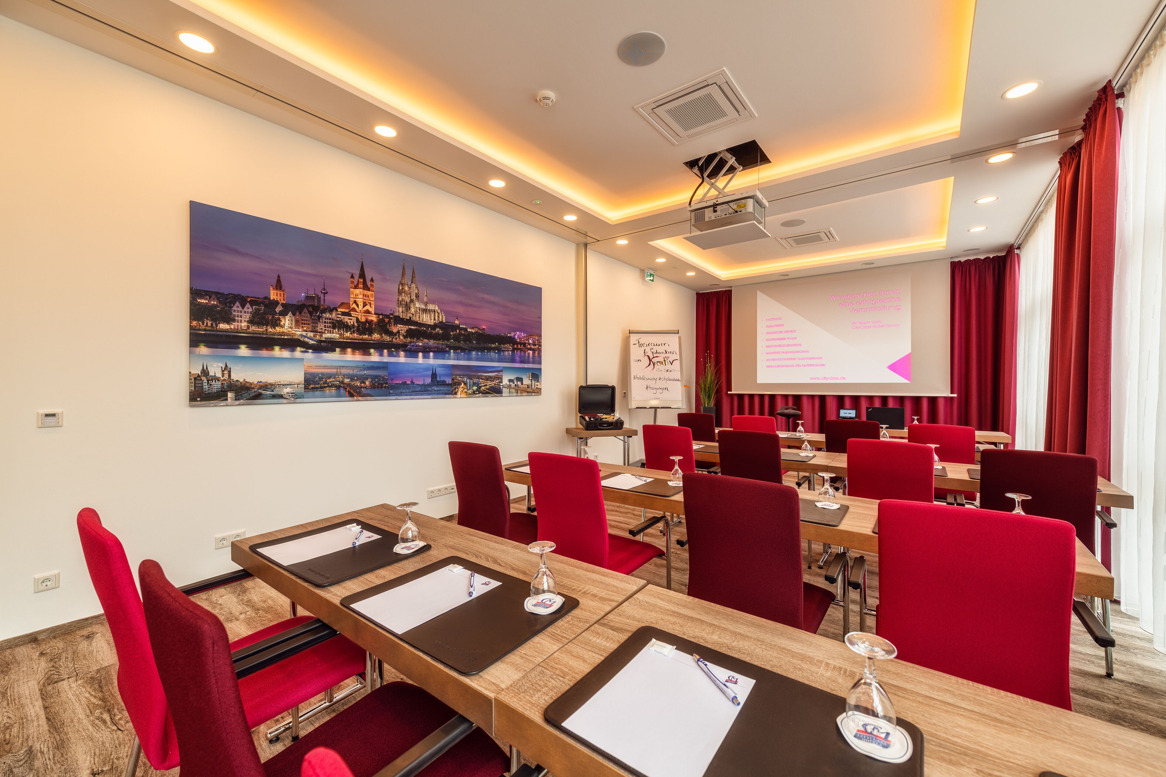 A conference room with red chairs, wooden tables, a projector screen, and a cityscape painting on the wall.