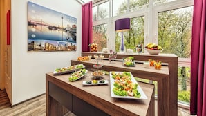 A dining area with a wooden table set with various dishes, a cityscape painting, and a view of trees through a window.