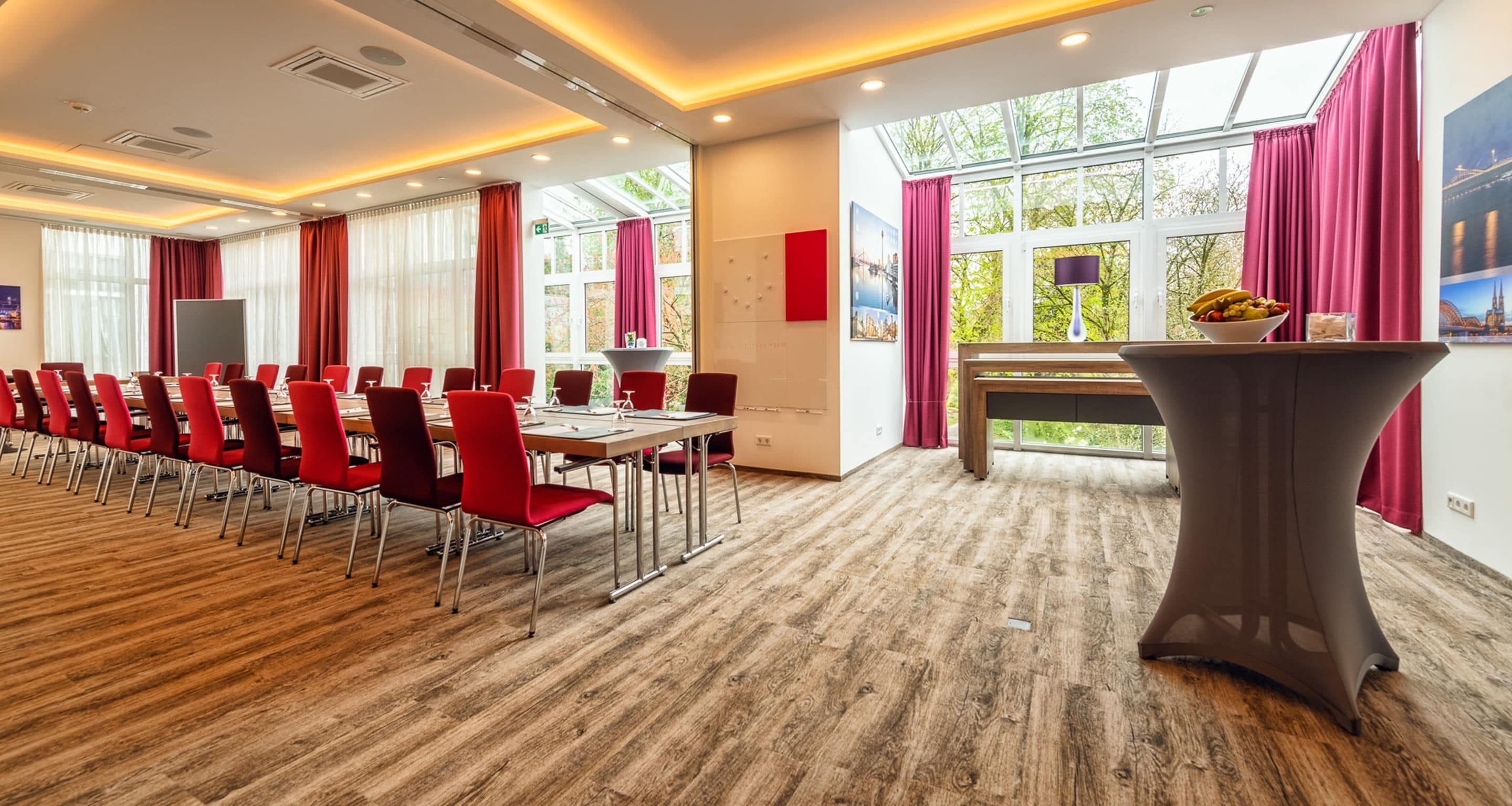 A conference room with a long table and red chairs, large windows with curtains, and a view of greenery outside.