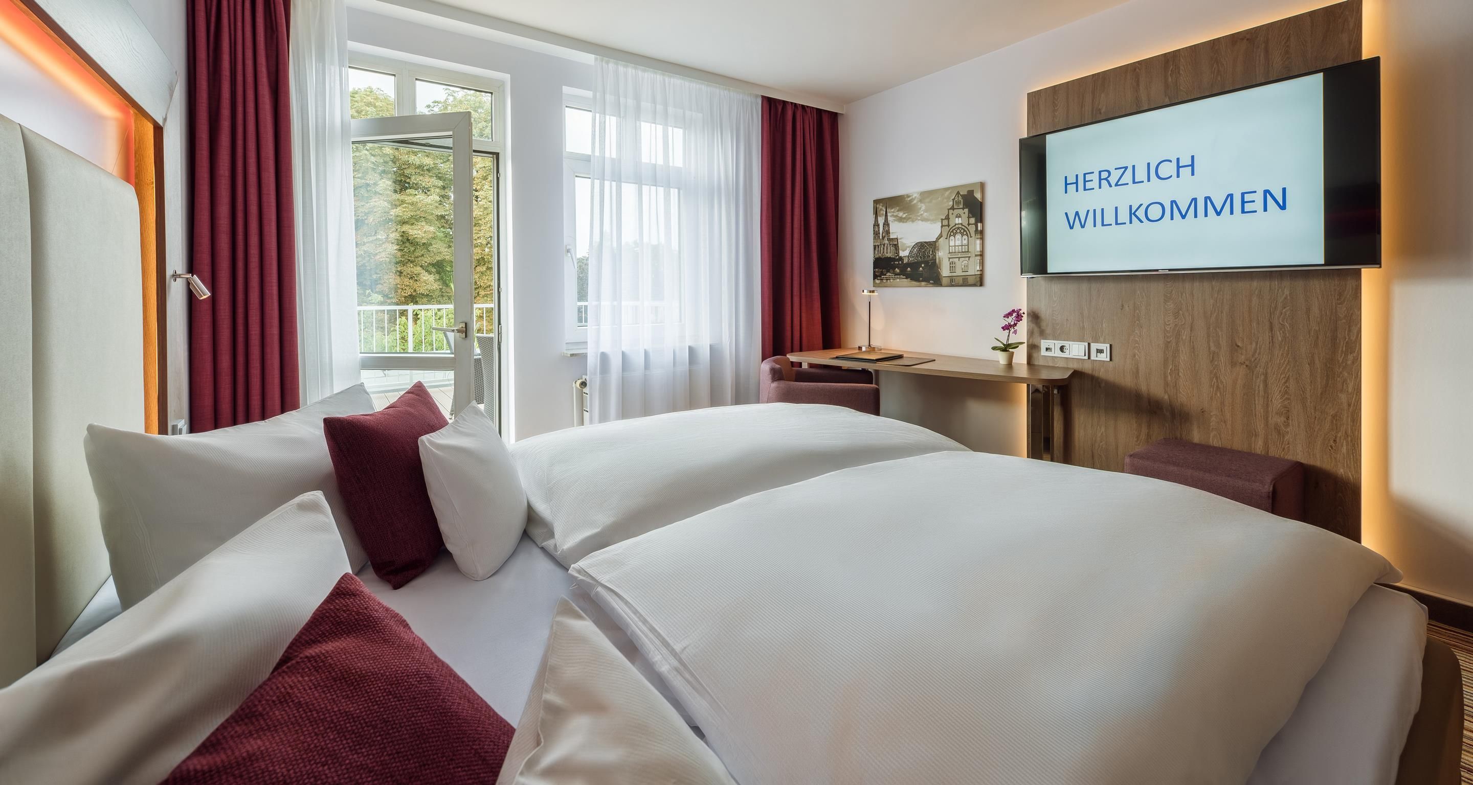 A hotel room with two beds, a desk, a TV, and a balcony with curtains.