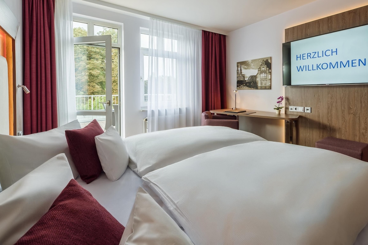 A hotel room with two beds, a desk, a TV, and a balcony with curtains.