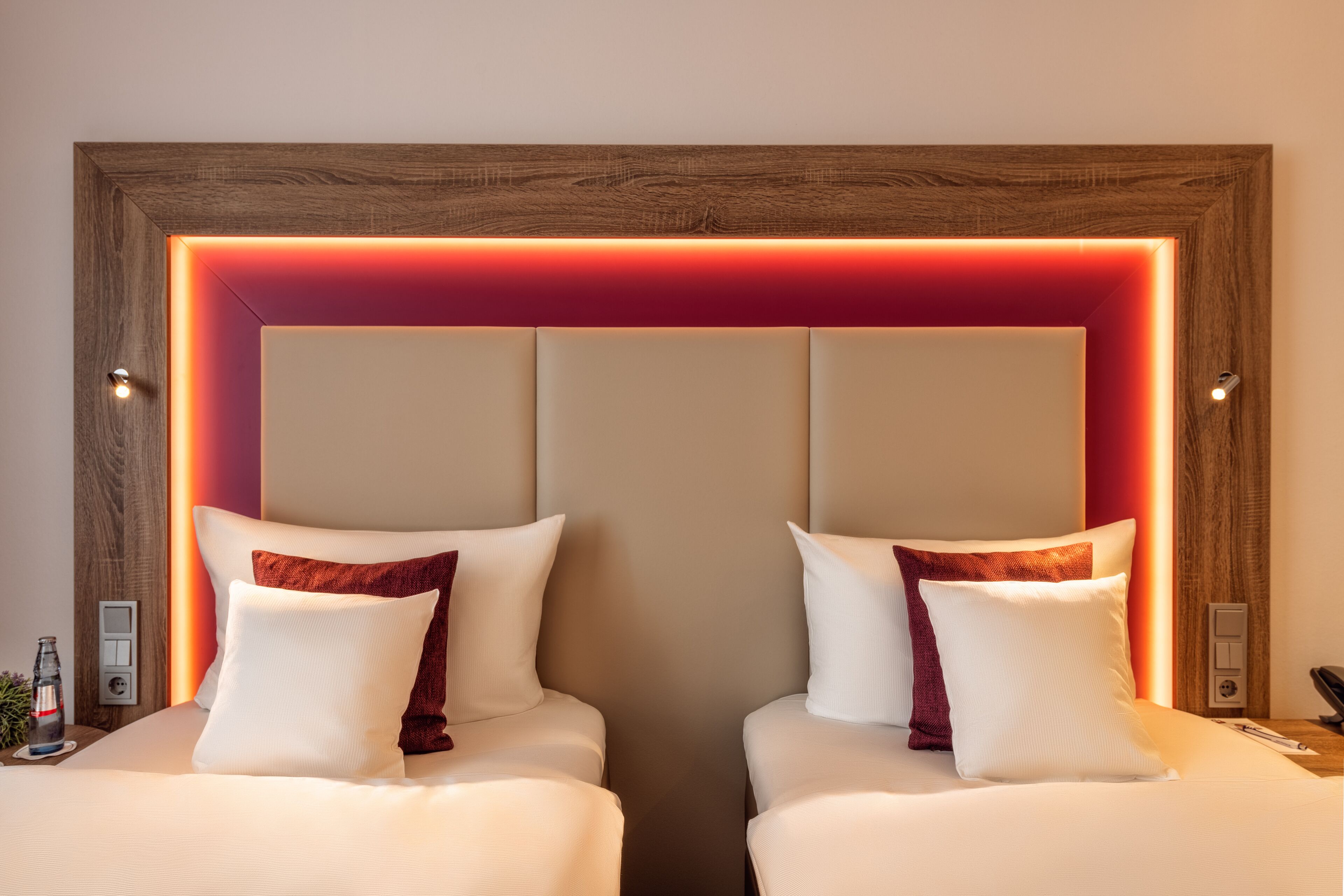 Habitación de hotel con dos camas, cada una con almohadas blancas y rojas, y cabeceros de madera con iluminación integrada.