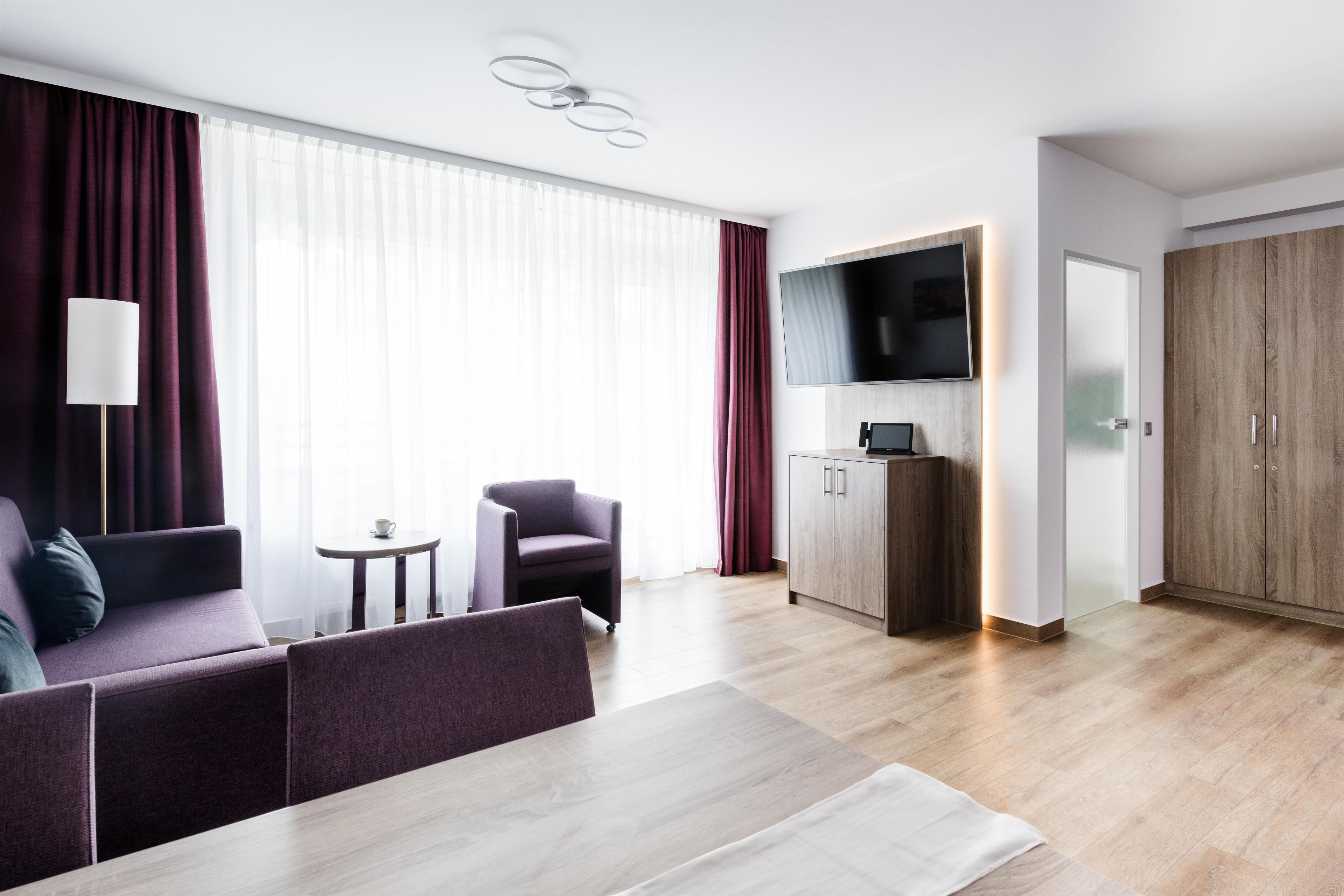 Una habitación de hotel moderna con sofá, sillón, mesa de comedor y televisor montado en la pared.