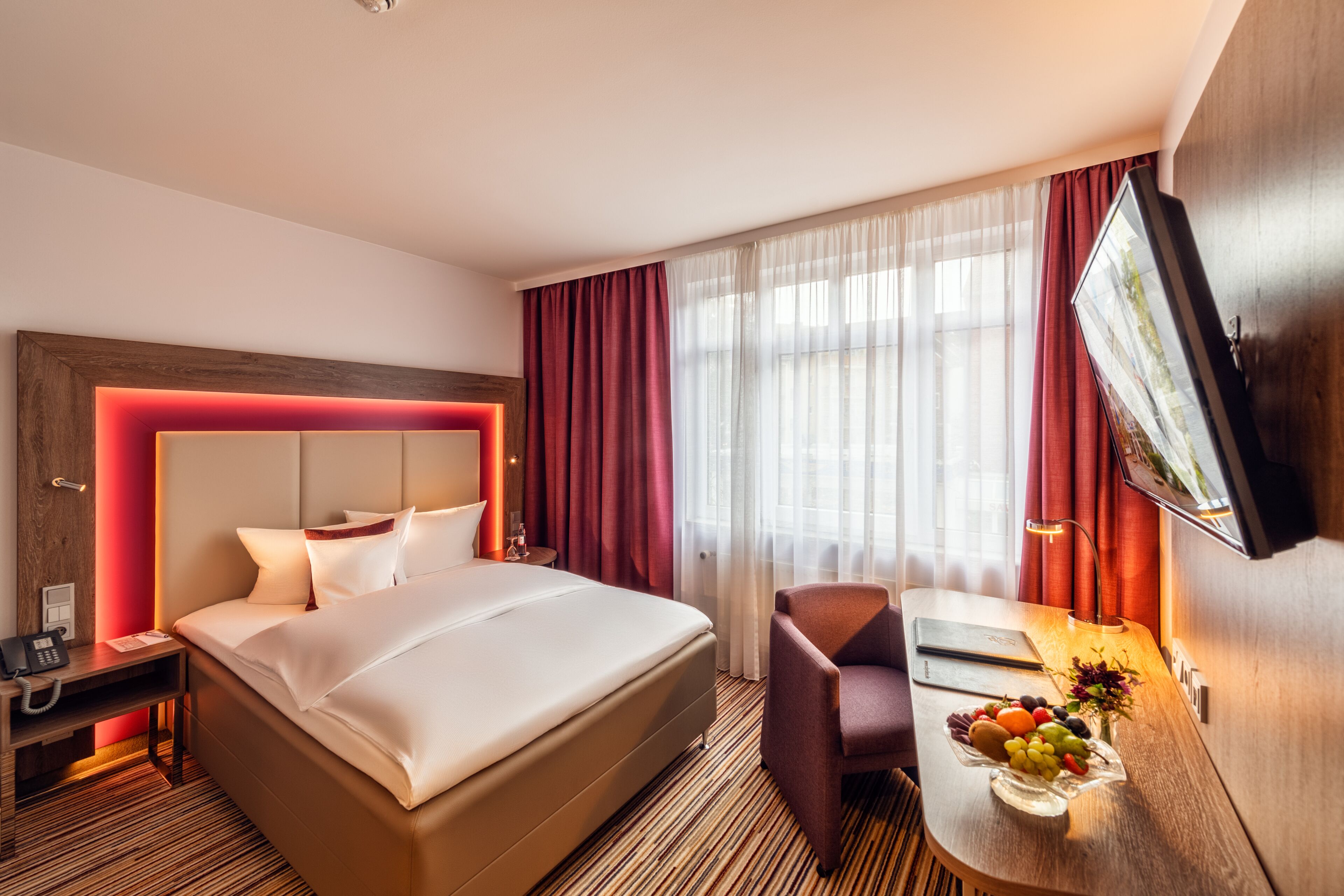 A hotel room with a large bed, a desk with a fruit bowl, a chair, a TV, and a window with curtains.