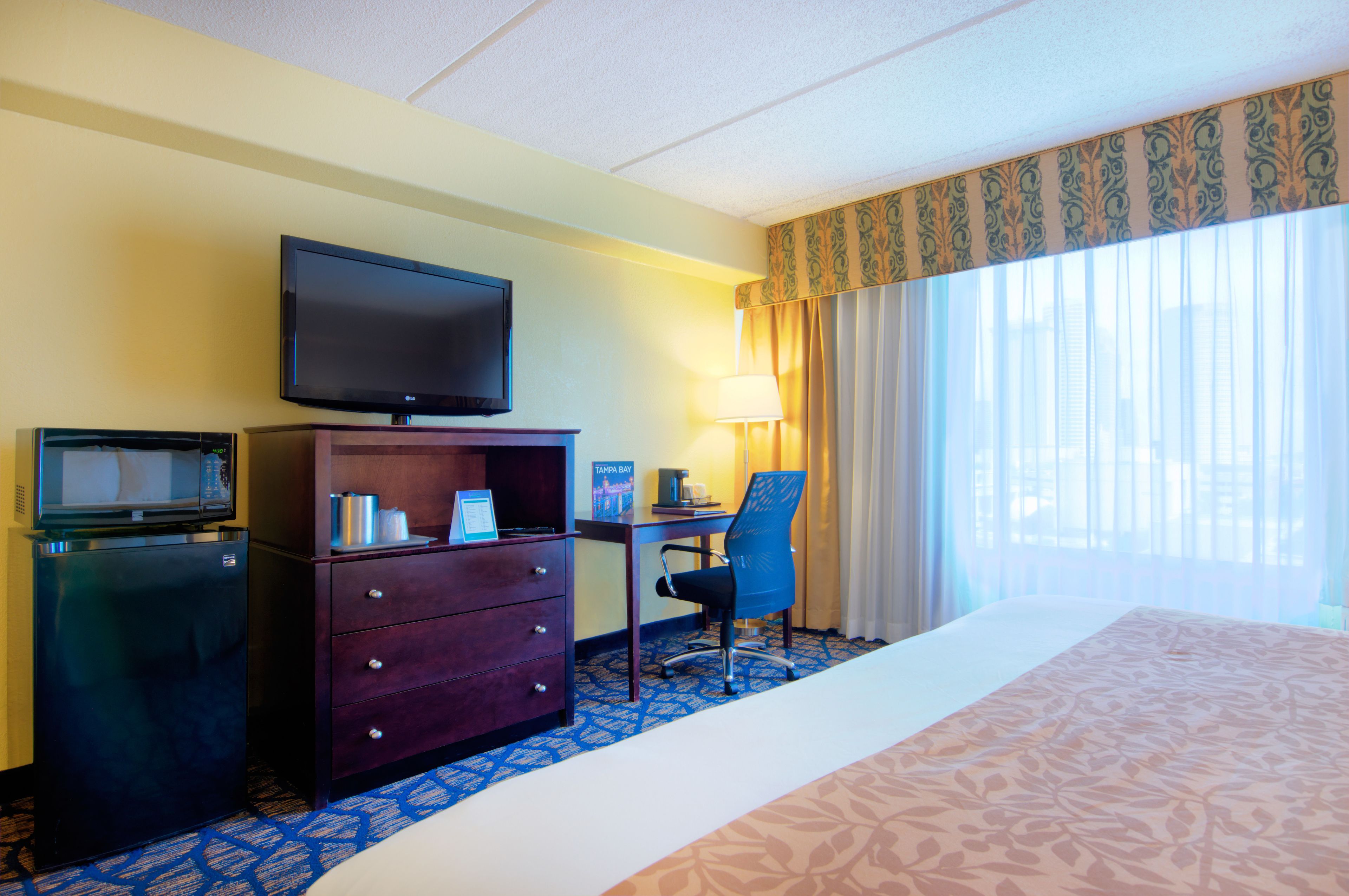 A hotel room with a bed, a desk with a chair, a television, a minibar, and a window with curtains.