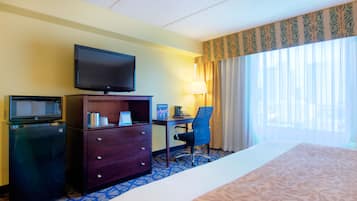 A hotel room with a bed, a desk with a chair, a television, a minibar, and a window with curtains.