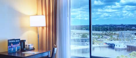 A hotel room with a large window offering a view of a cityscape, a desk with a lamp, and a coffee maker on the table.